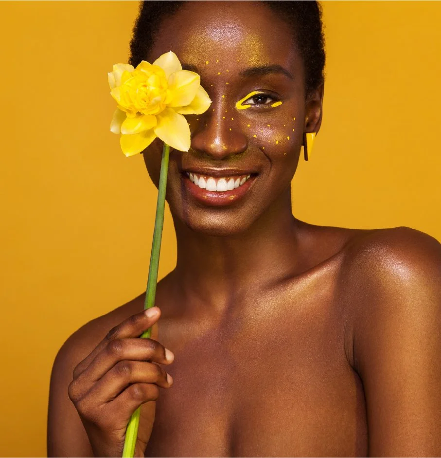 A smiling woman with dark skin holding a yellow flower in front of one eye against a yellow background.