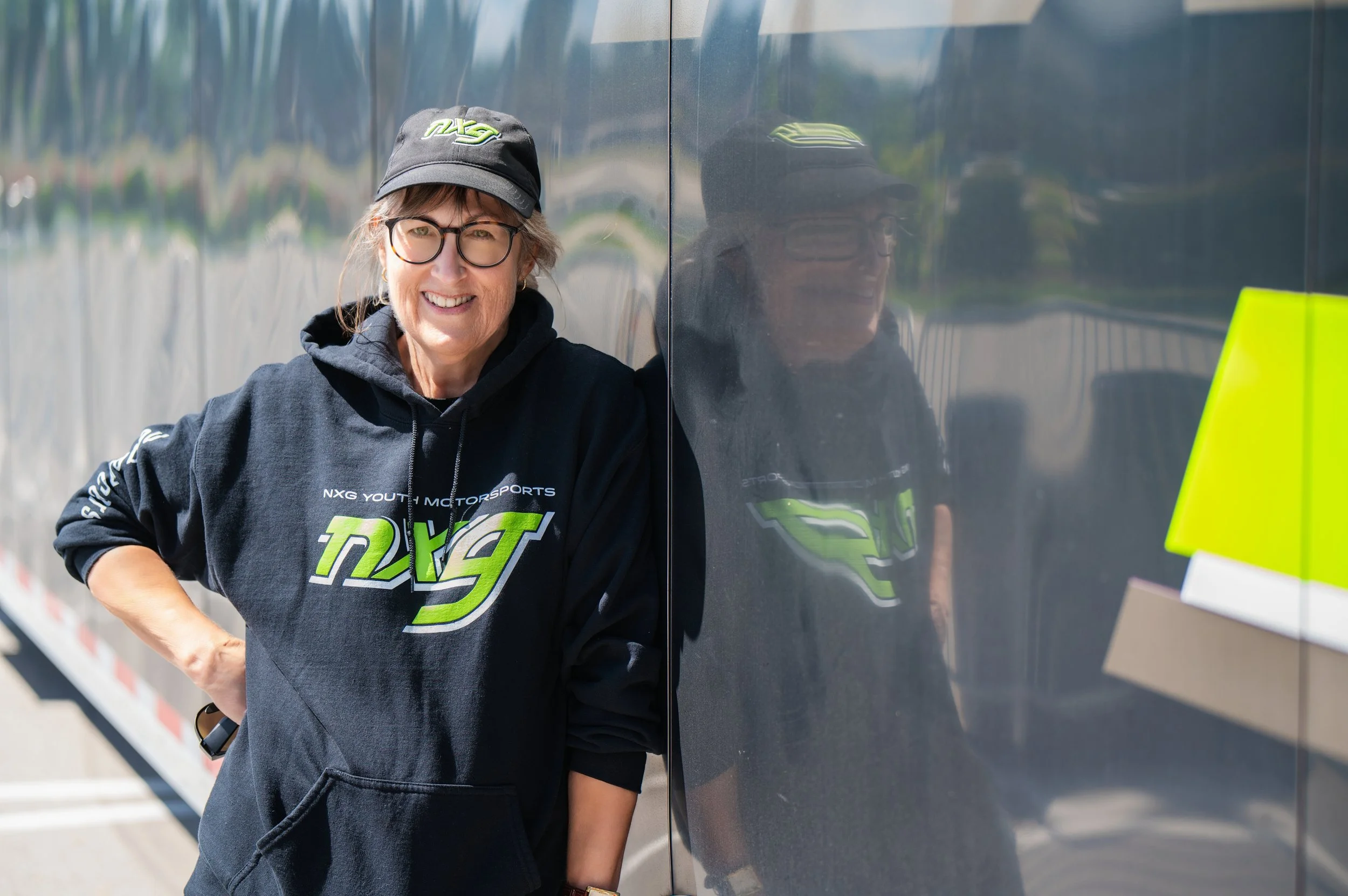 Person wearing a black NXG Youth Motorsports hoodie and cap standing outdoors next to a reflective surface.