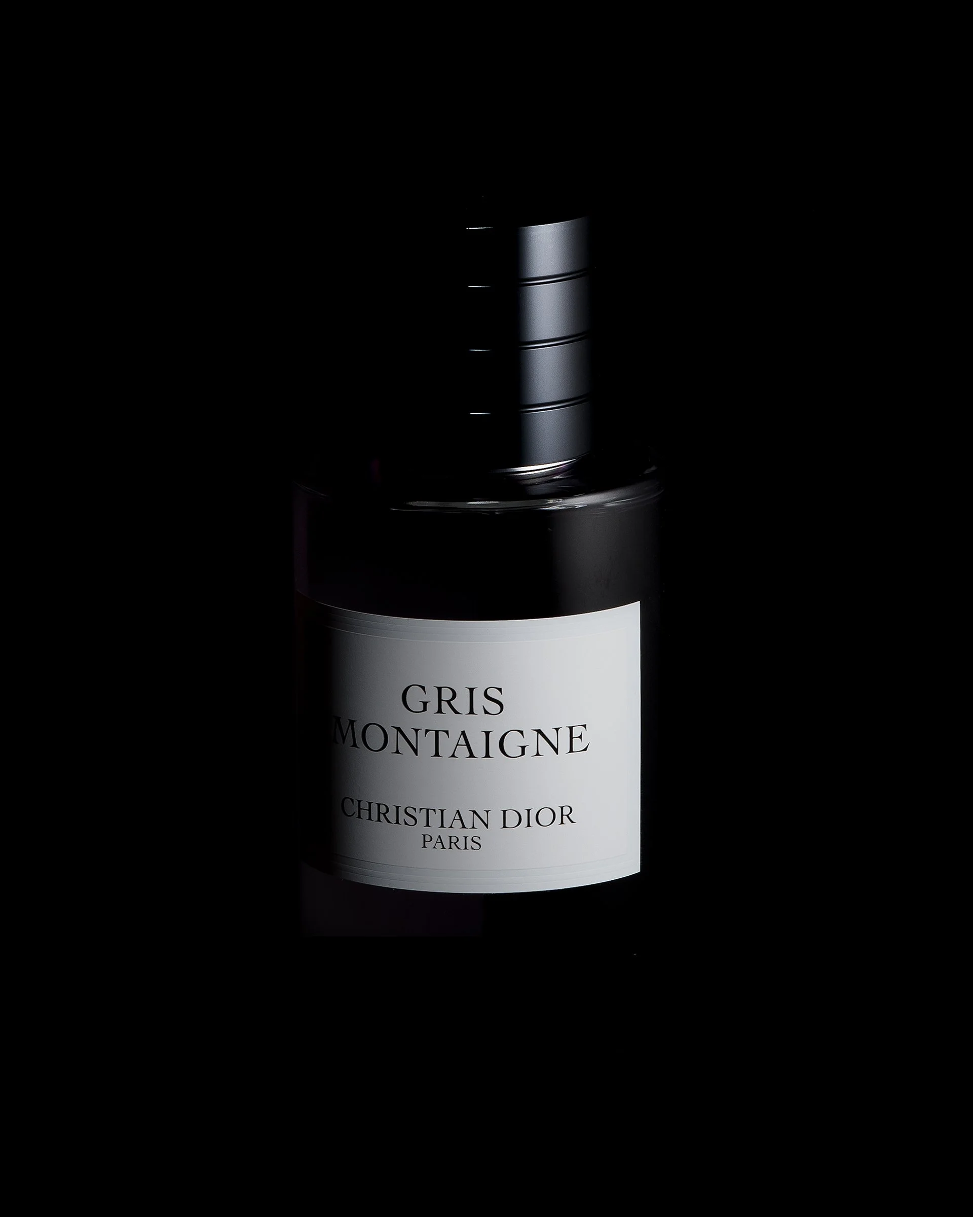 A dark background with a partially illuminated bottle of Christian Dior Gris Montagne perfume. The label is visible, displaying the perfume's name. Photography by Kenneth Foo.