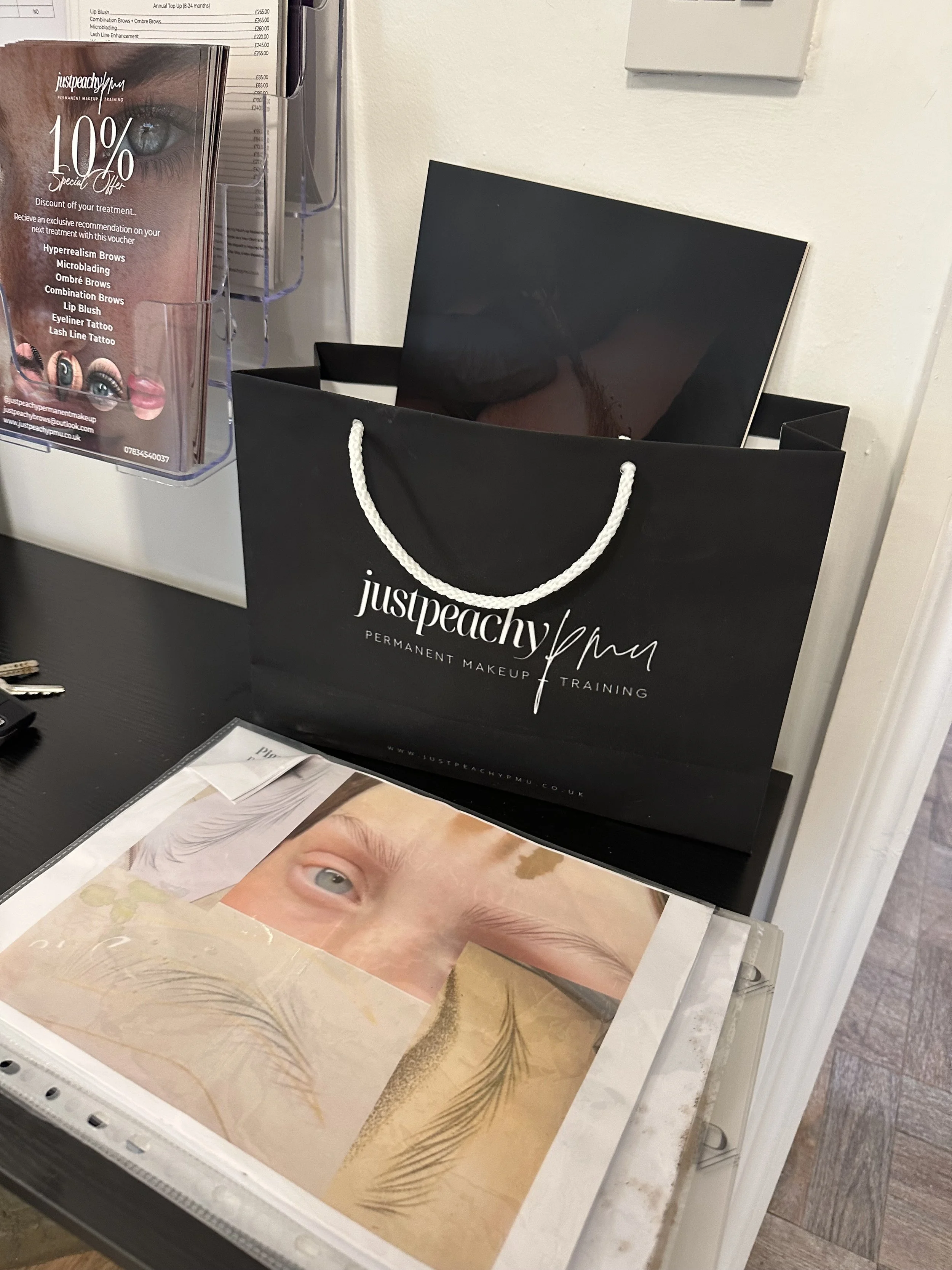 A black with white text shopping bag for 'justpeachypmu' permanent makeup training, a laminated poster showing an eye and eyebrows, a brochure stand with promotional materials, keys on a keyring, and a glass display with eyelash images.