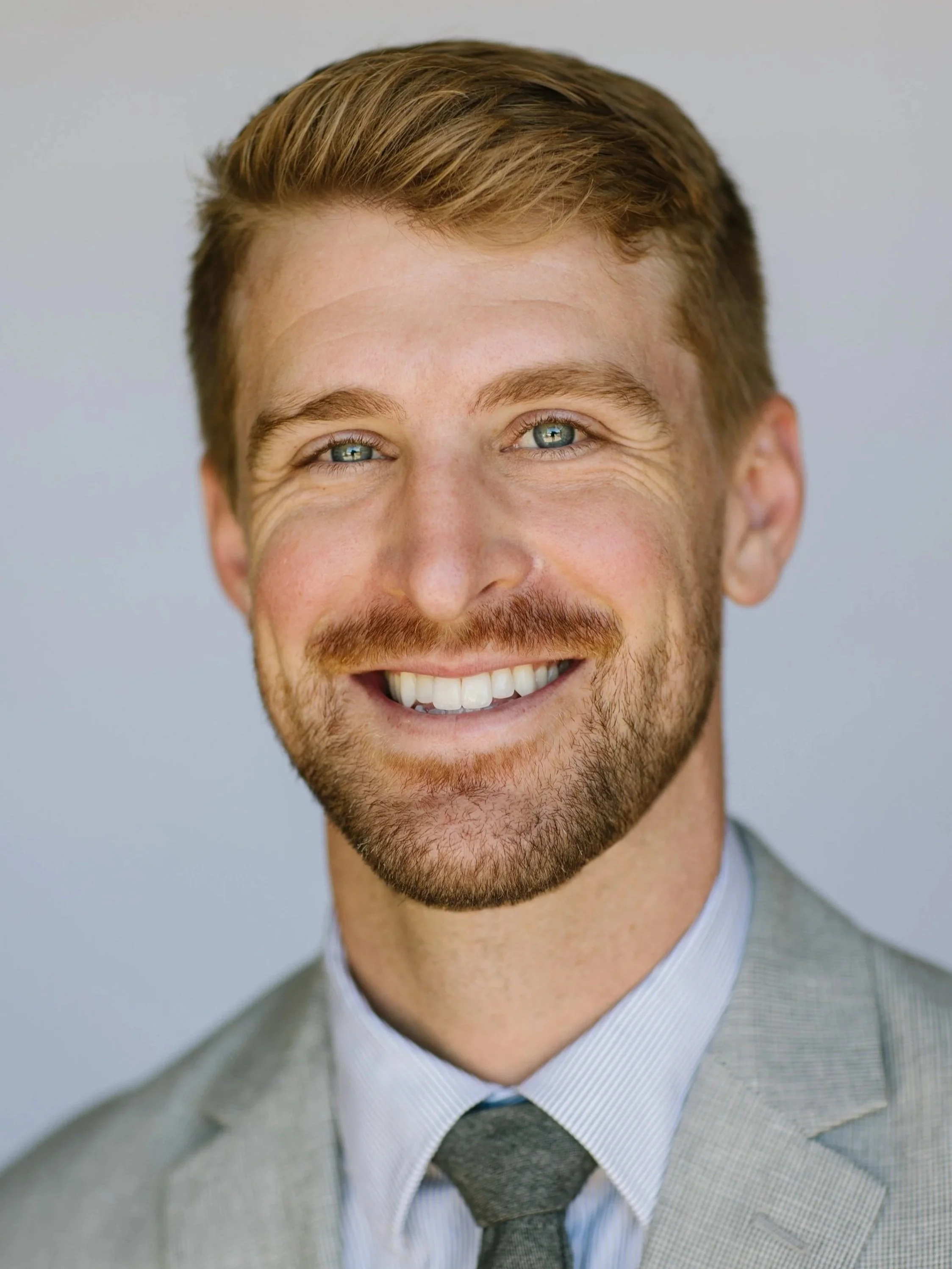 A smiling man in a light gray suit with a checkered pattern, white collared shirt, and dark tie, featuring short light brown hair, blue eyes, and a light complexion.