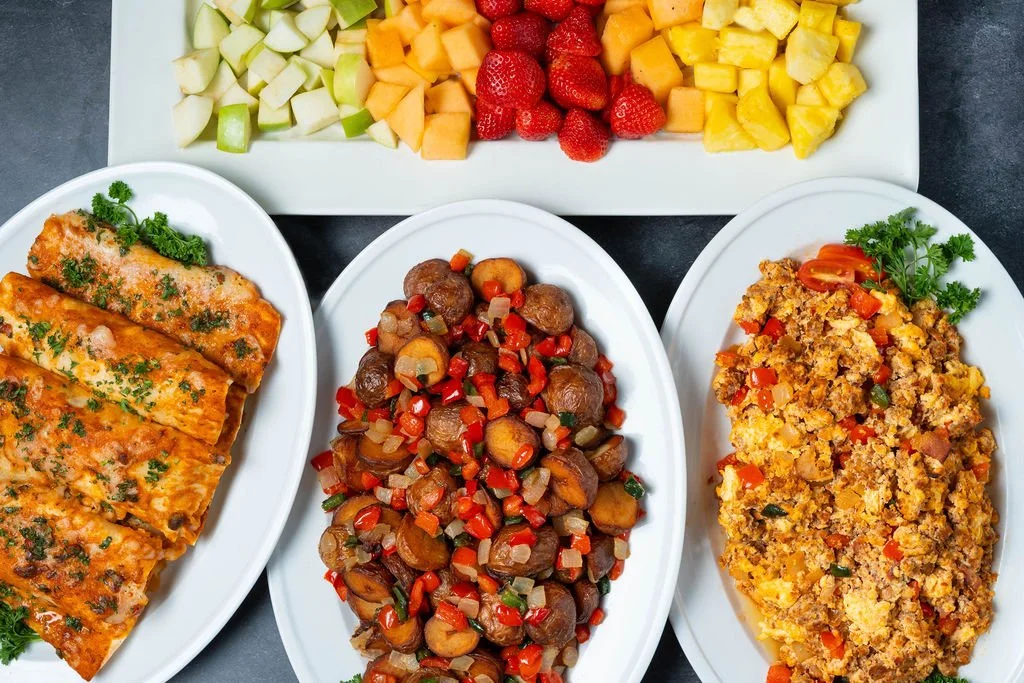 Catering spread with three large platters of prepared dishes and a tray of cut fruit, arranged on a table for a catered meal.