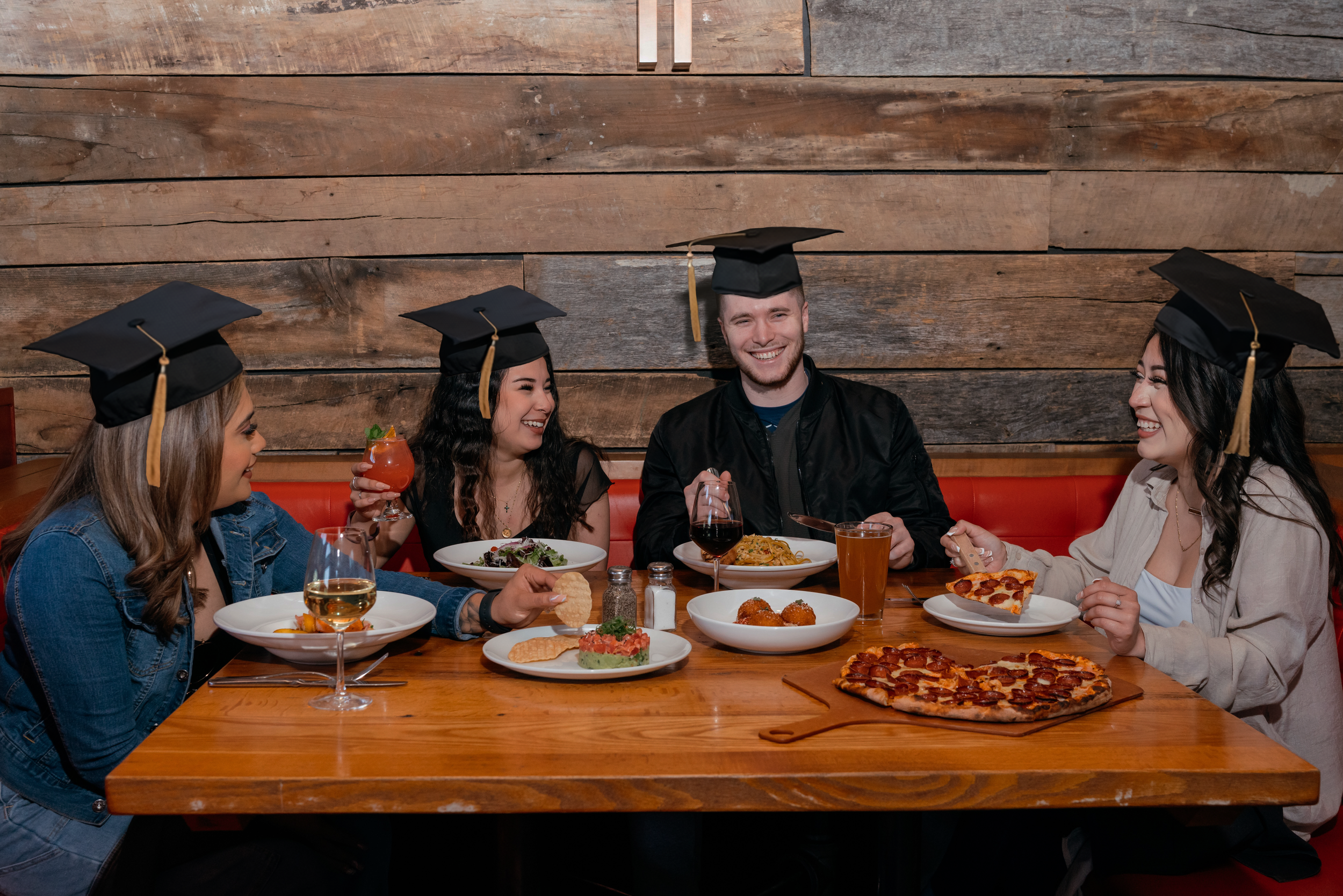 Group of adults wearing graduation caps seated at a restaurant table, sharing pizza, entrées, and drinks in a casual dining setting.