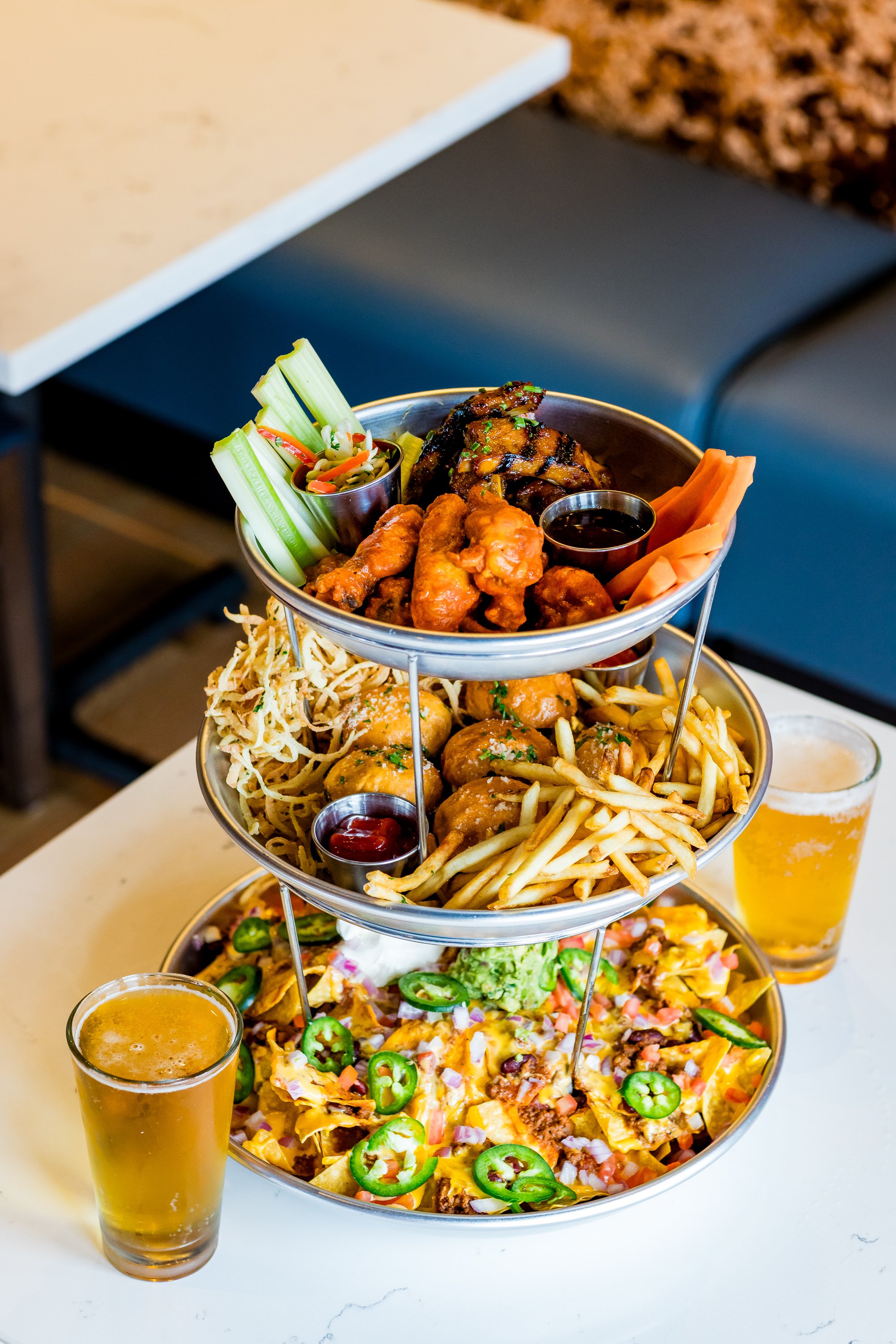 A three‑tier tray filled with wings, grilled chicken, fries, fried snacks, and loaded nachos, with two glasses of light beer on a white table in a casual dining setting.