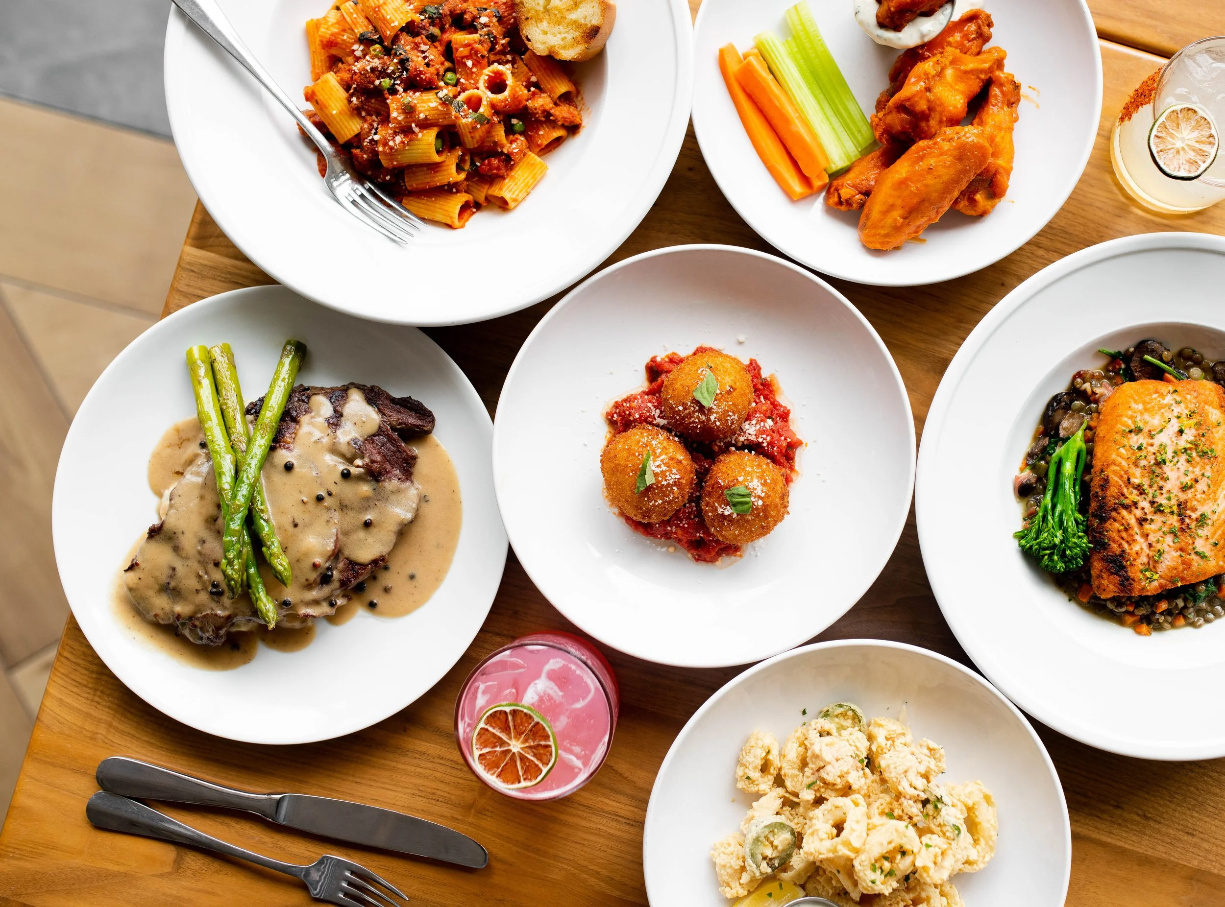 Overhead view of a wooden table with multiple plated dishes, including pasta, meat entrées, wings, vegetables, and a cocktail arranged for sharing.