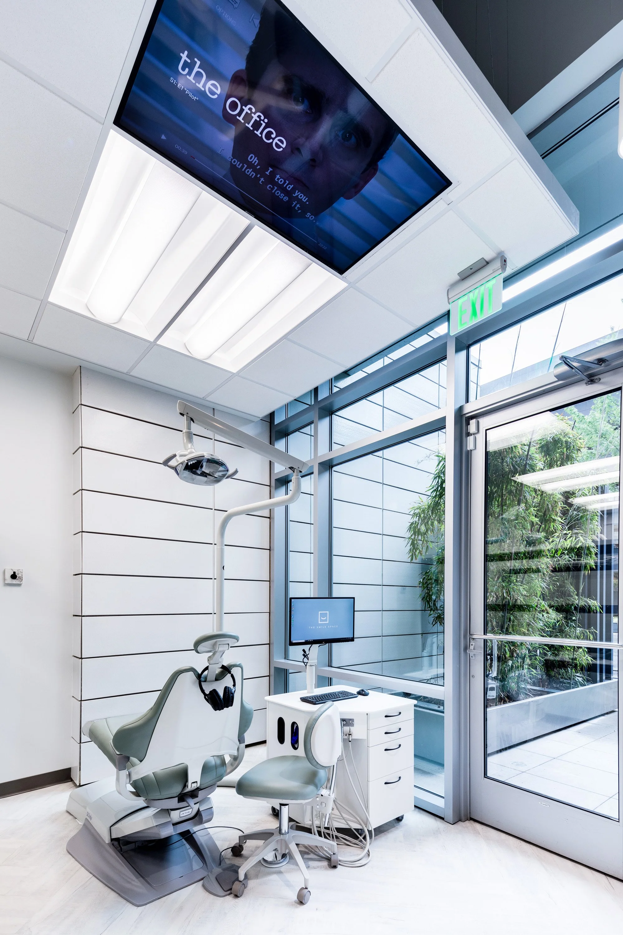 Modern dental treatment room at The Smile Space featuring floor-to-ceiling windows, a ceiling-mounted TV, and a comfortable patient chair.