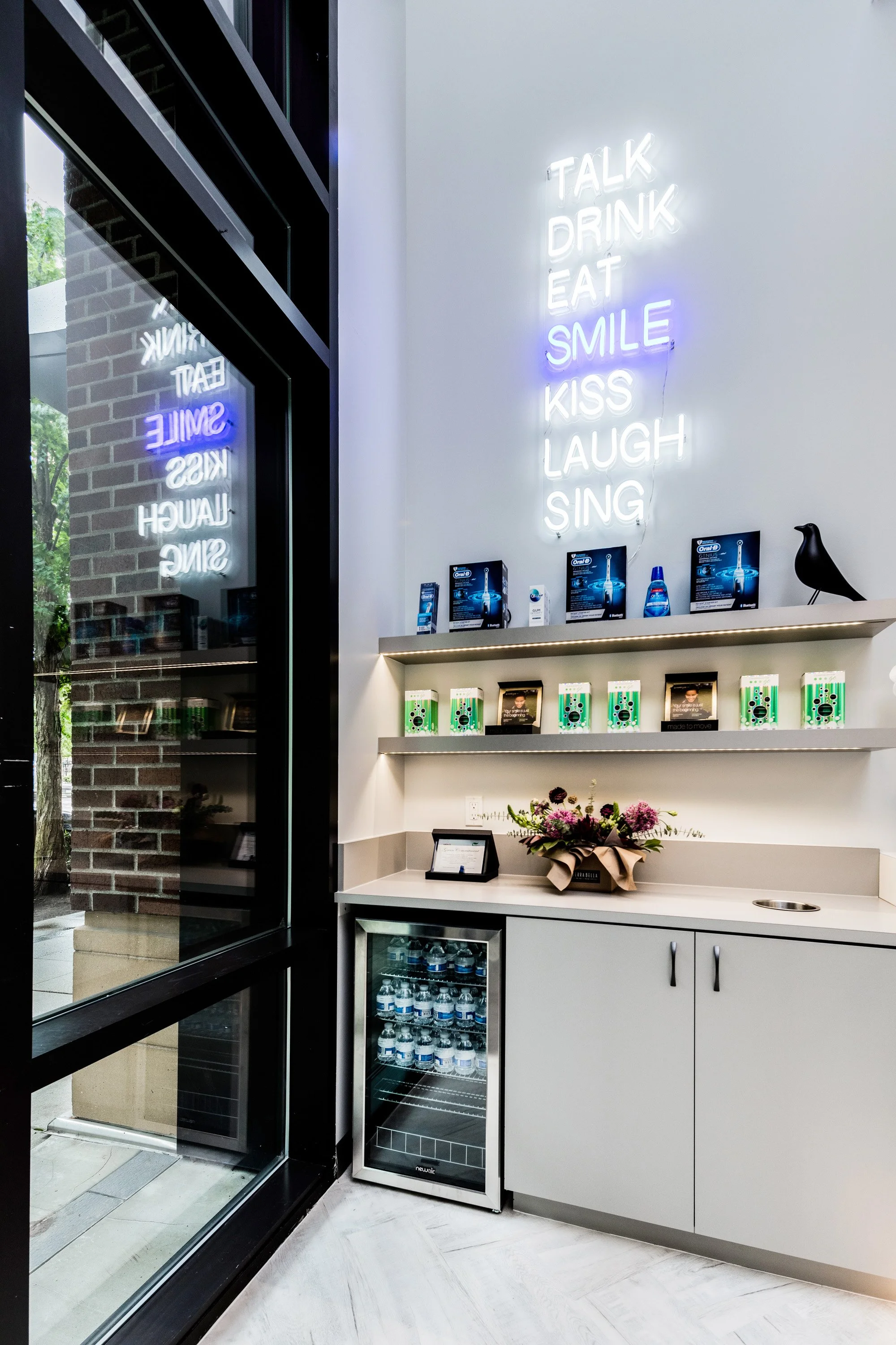 Patient refreshment area at The Smile Space featuring a neon "Talk Drink Eat Smile Kiss Laugh Sing" sign, complimentary beverages, and oral care products.