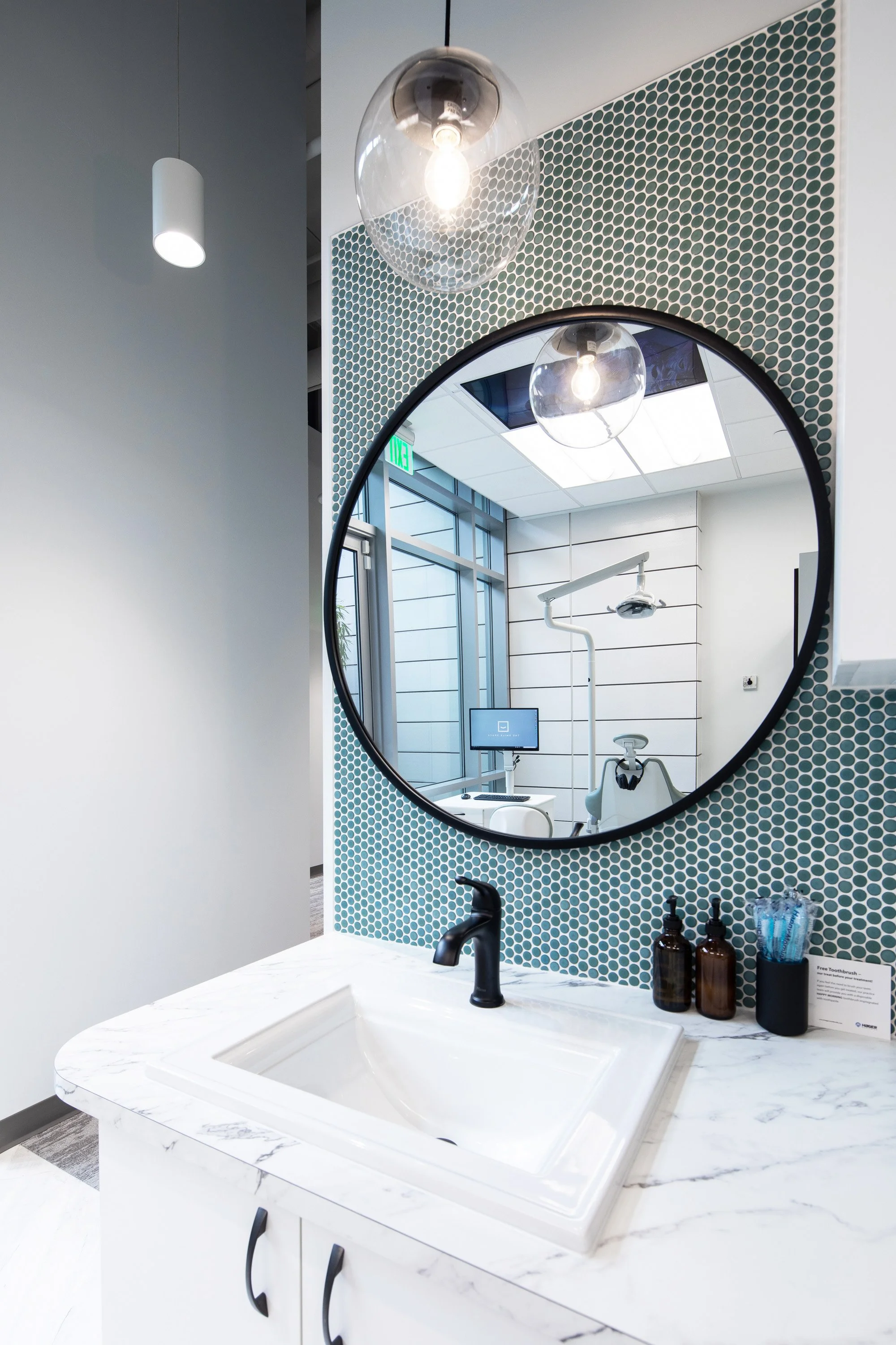 Patient brushing station at The Smile Space featuring a sink, marble countertop, round mirror, and complimentary toothbrushes before dental treatment.