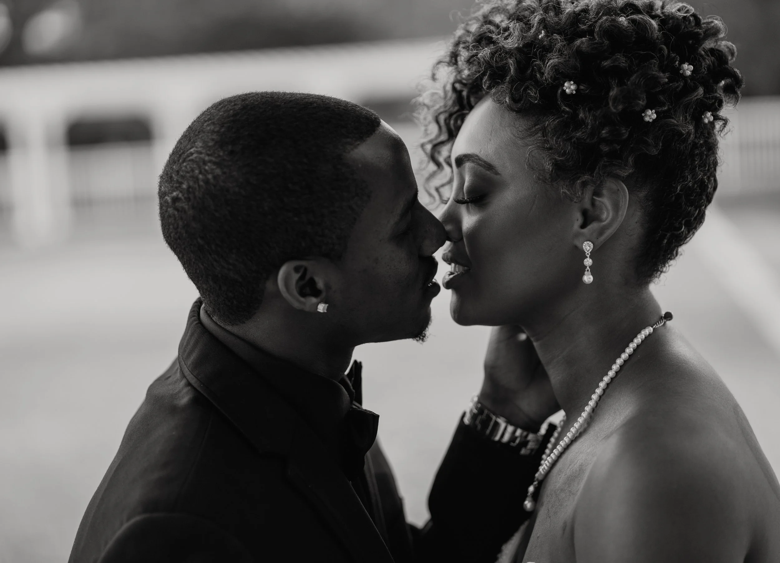 A black and white photograph of a romantic moment between a man and a woman about to kiss, with the woman wearing jewelry and having curly hair and the man having short hair and earrings.