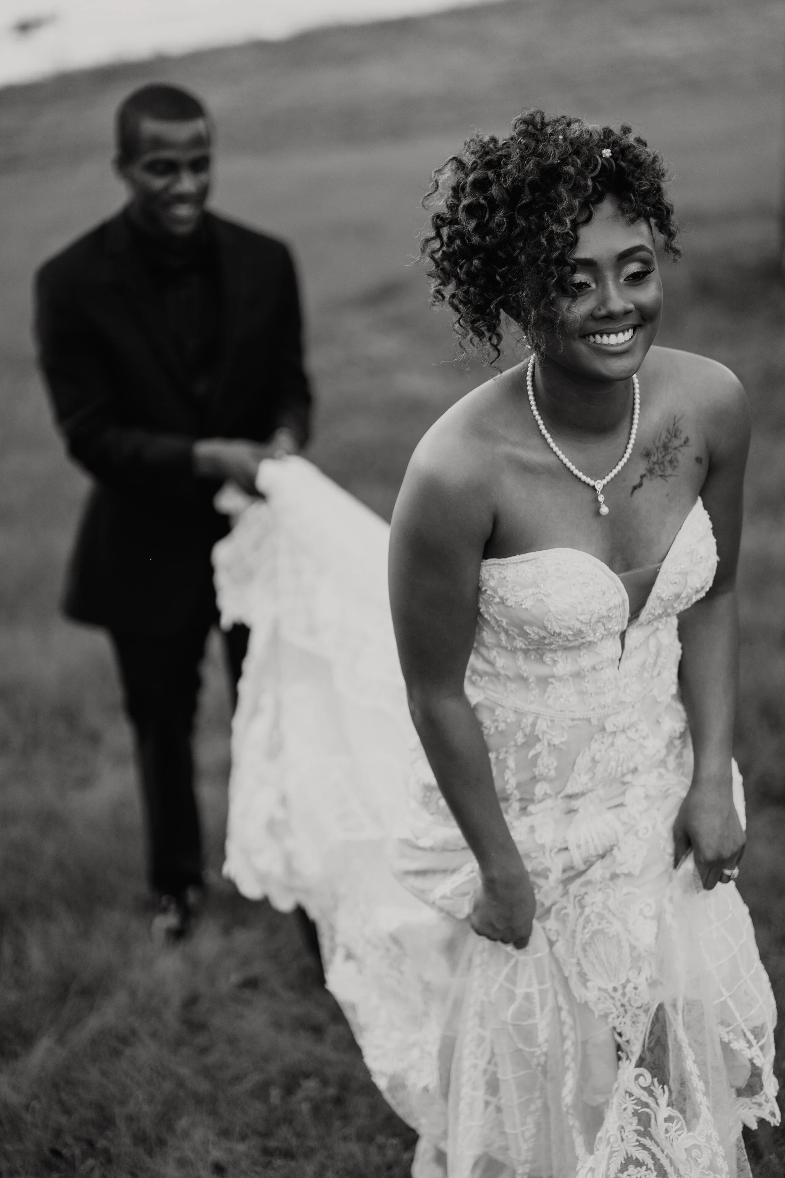 A black-and-white photo of a bride smiling in her wedding dress, holding the bottom of her dress, with a groom in a suit smiling in the background, holding a cloth or garment.