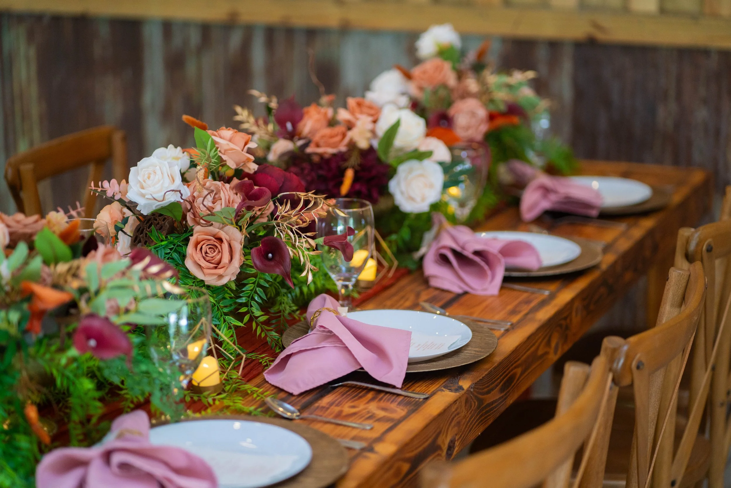 A long wooden dining table set for a special occasion with pink napkins and white plates, decorated with a lush floral centerpiece of roses, calla lilies, and greenery, with candles.