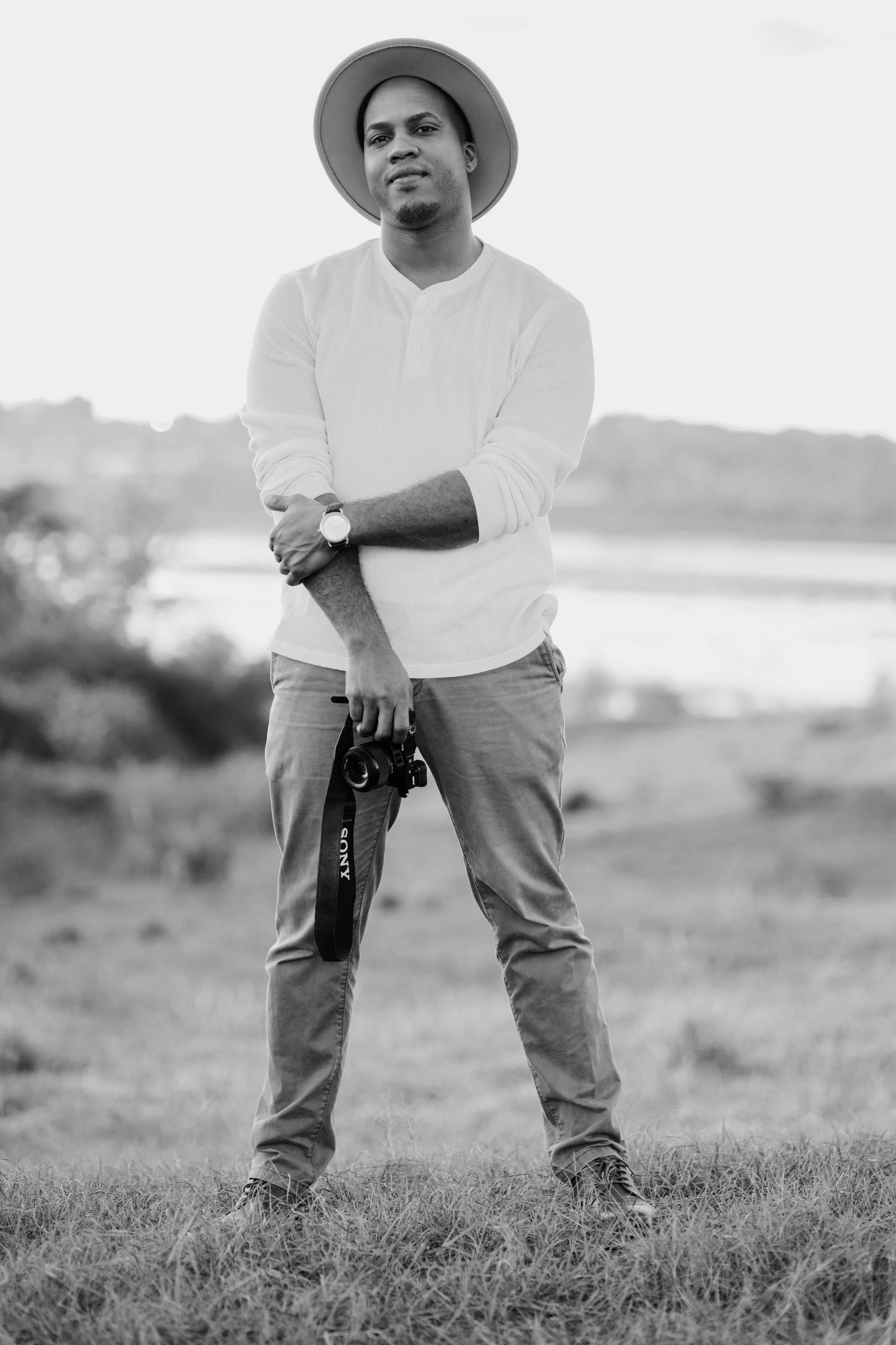 fresh creativo  wearing a wide-brimmed hat and casual clothing, holding a camera in one hand, standing outdoors in a grassy area with a blurred landscape in the background.
