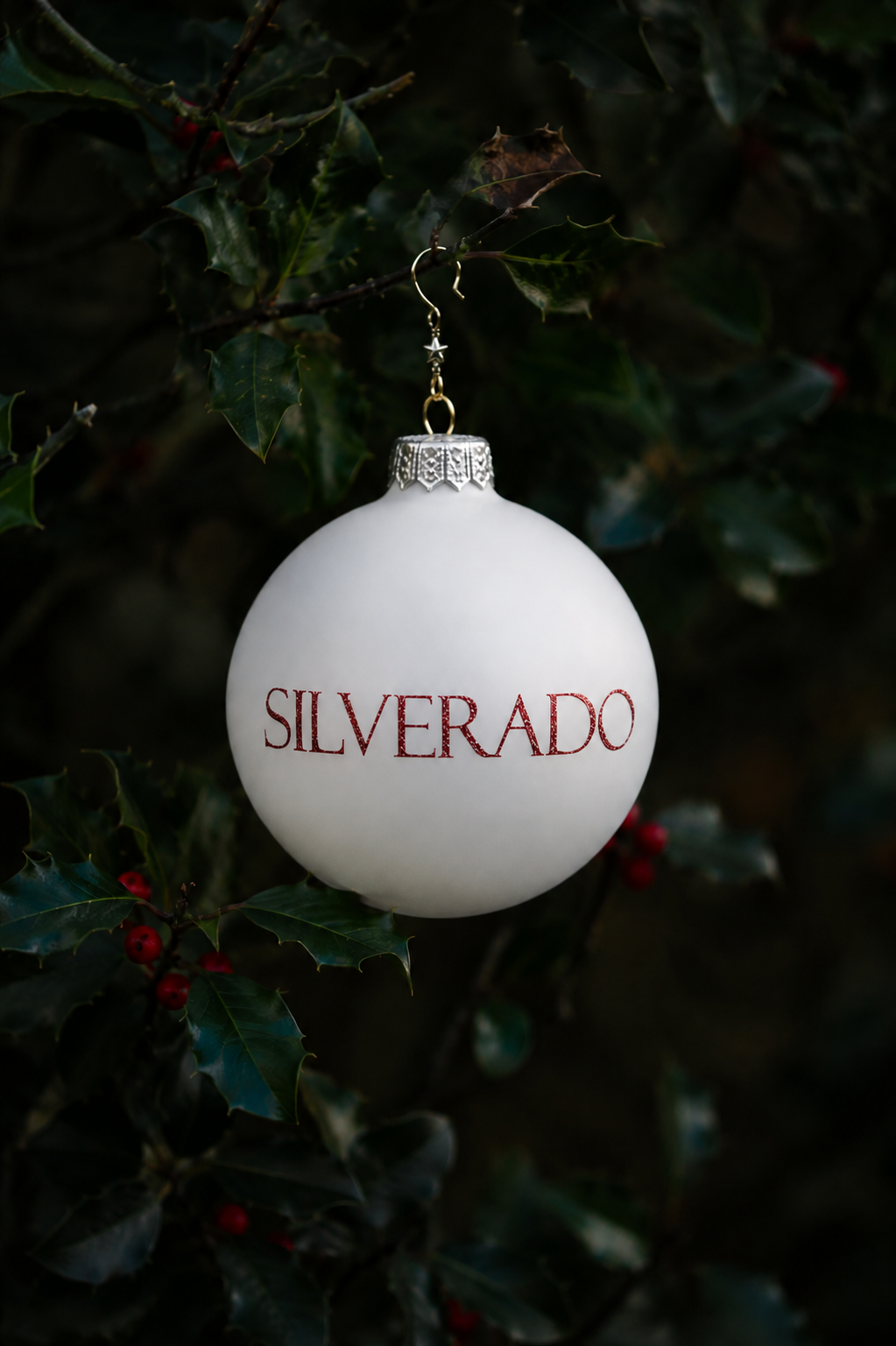 A white Christmas ornament hanging on holly branches with dark green leaves and red berries, with the word "SILVERADO" written in red on the ornament.