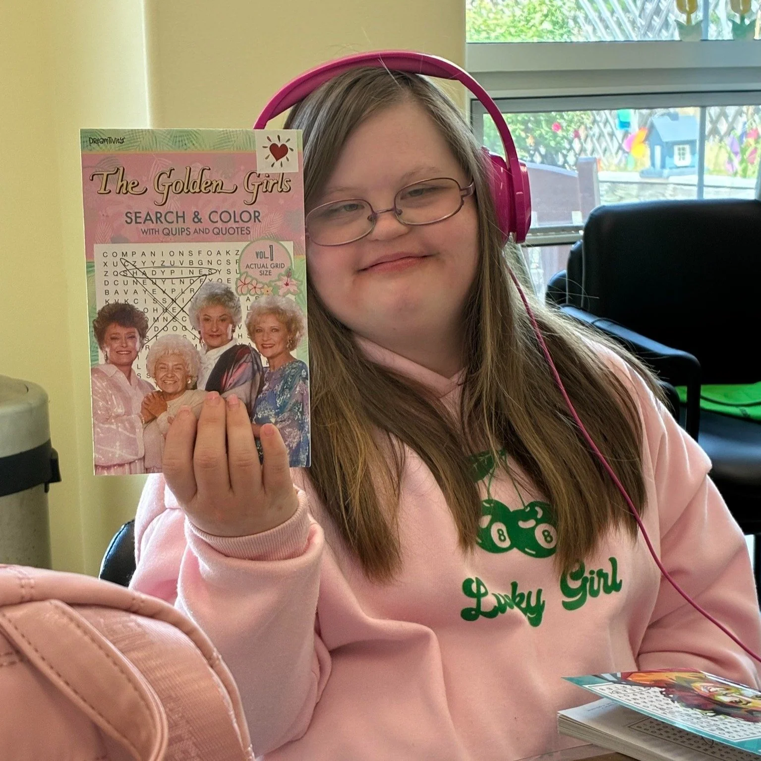 woman in her 20s wearing pink headphones holding up a golden girls word search book