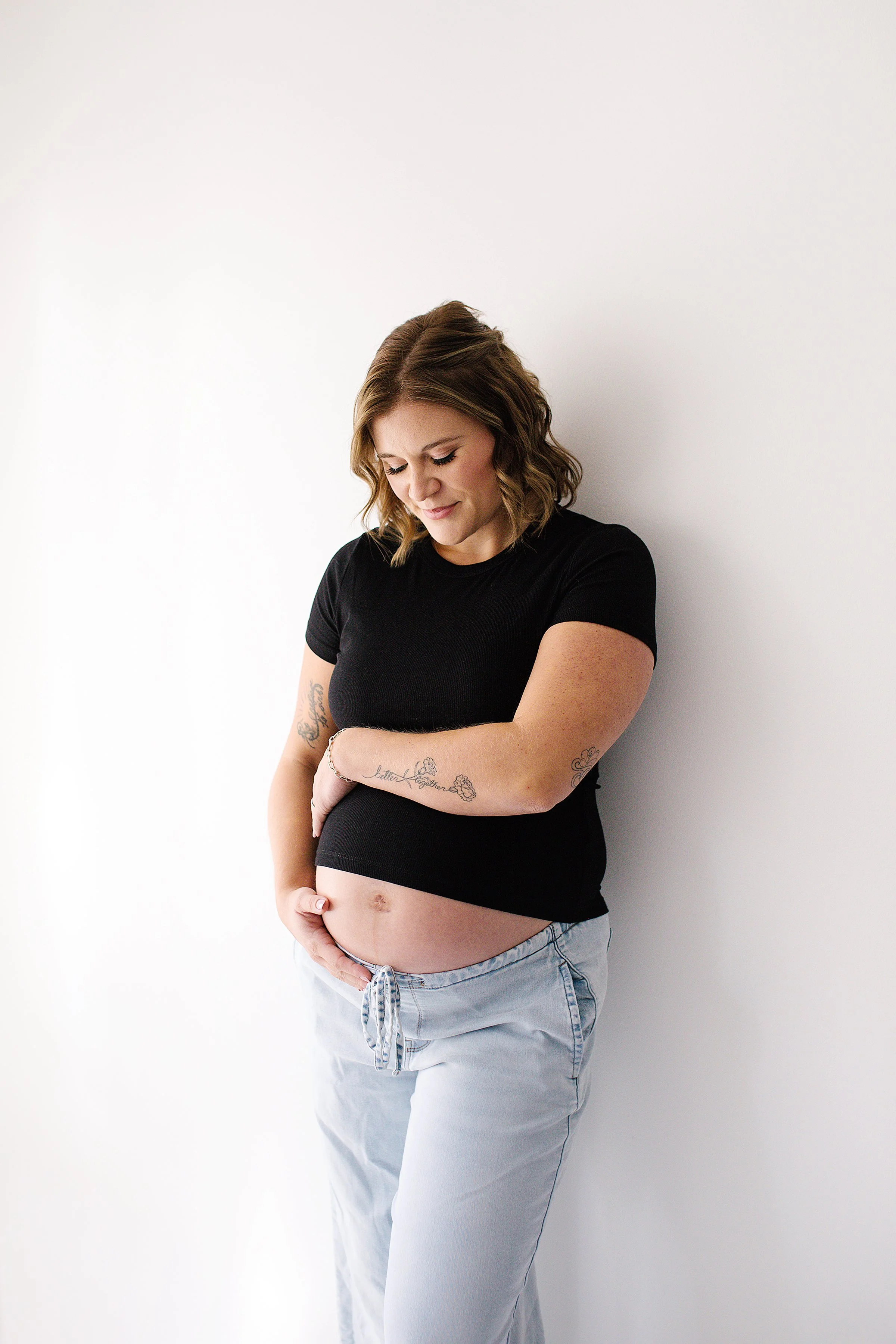 Pregnant mom leaning on wall in black cropped tee and jeans with hands on belly