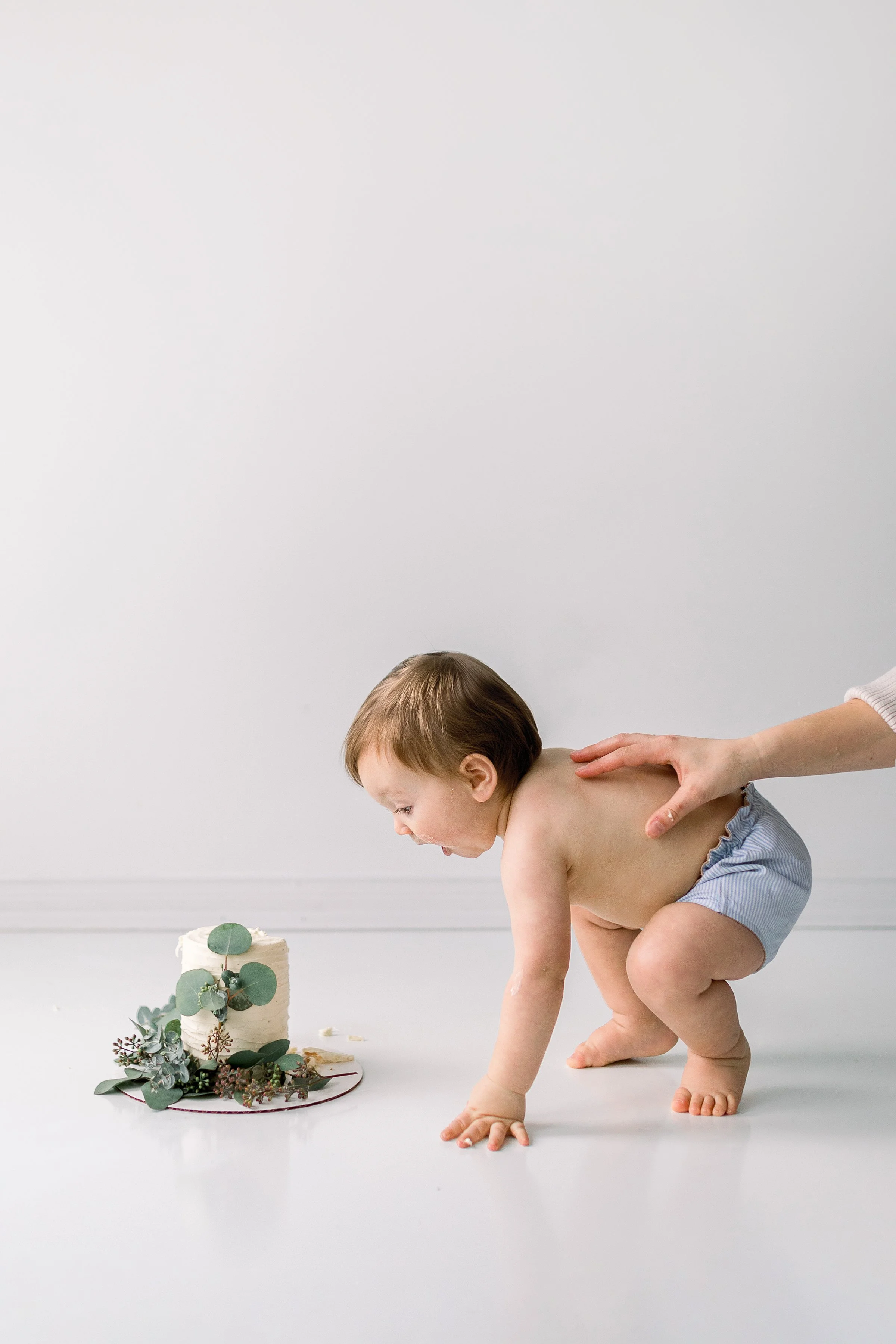 Cute baby boy leaning down to tea e bite of his floral cake smash with mom holding onto his back, in Durham Region