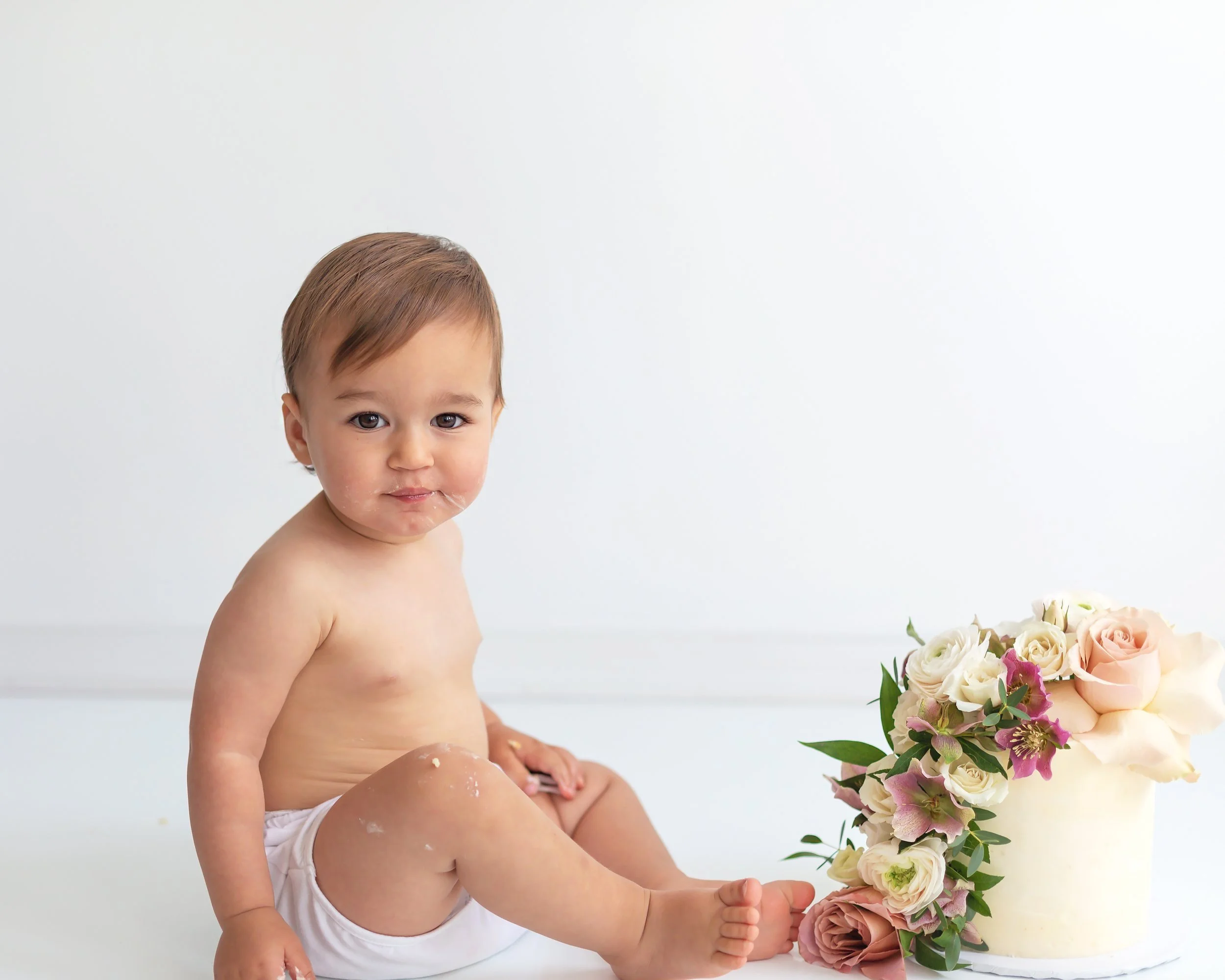 Cute baby girl in white diaper cover sitting facing her gorgeous floral cake smiling at camera, in Oshawa baby studio