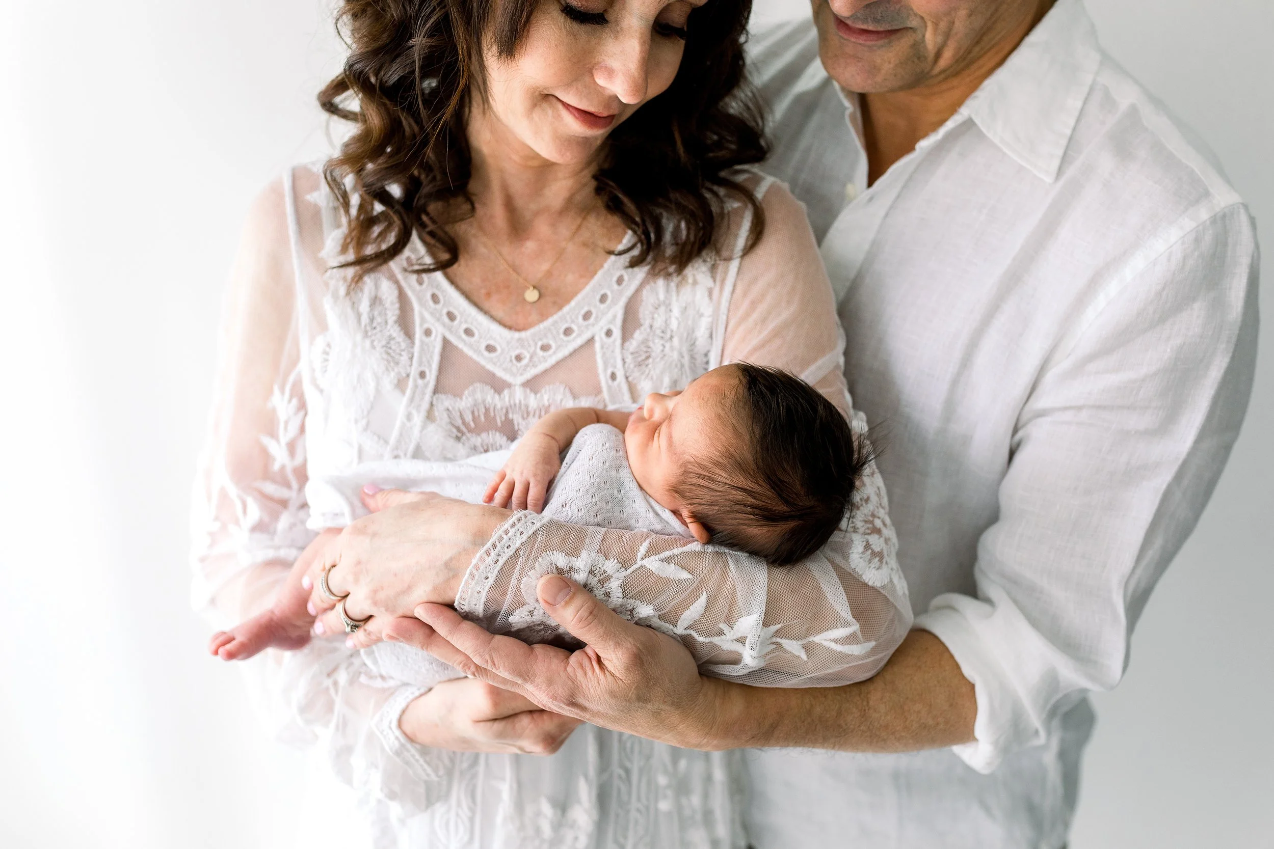 New parents holding their newborn baby girl in a white lace dress and linen shirt, smiling at her in an Oshawa studio