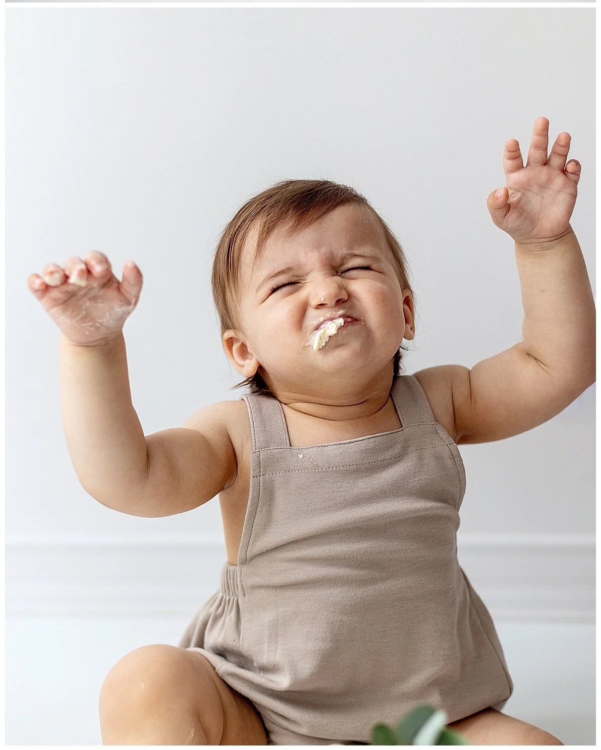 Cute one year old baby boy in brown romper with a piece of icing in his mouth squinting and hands up, in Oshawa studio