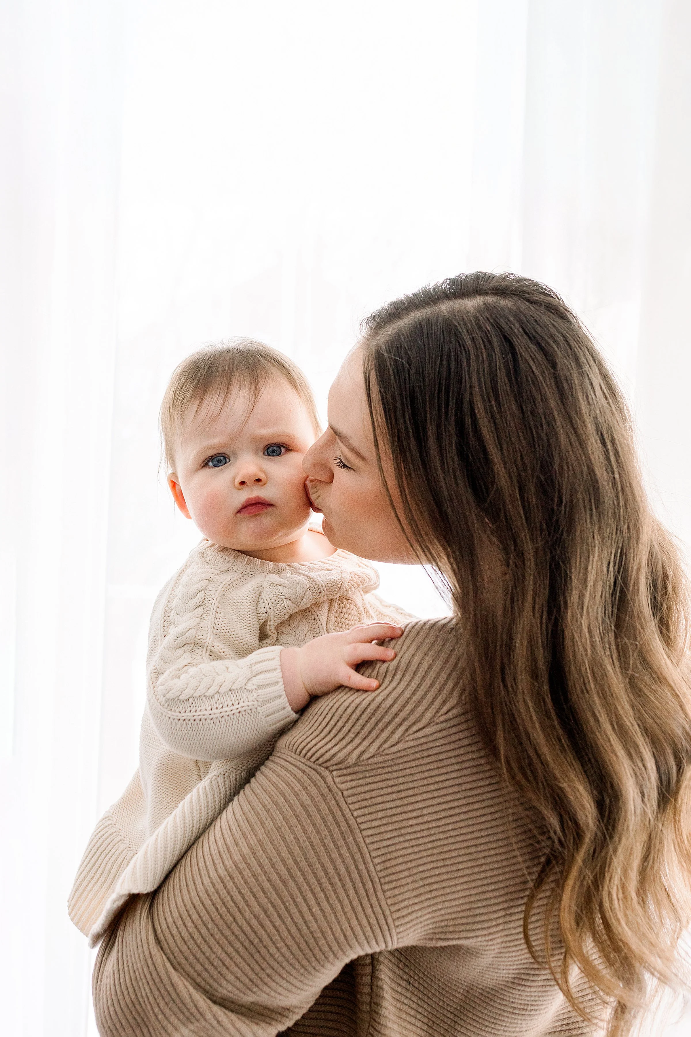 A woman holding a baby in front of a white curtain, with the woman kissing the baby's cheek.