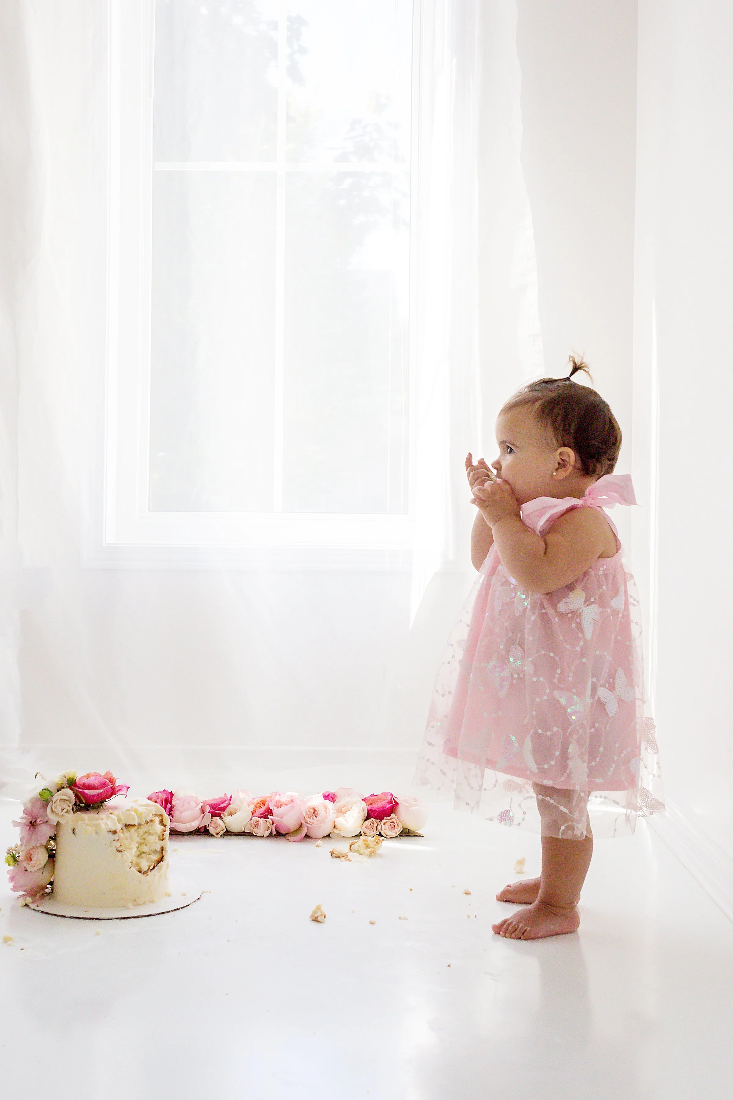 One year old baby girl in pink lace dress standing up eating a piece of her pink floral cake in front of a white window, in Oshawa