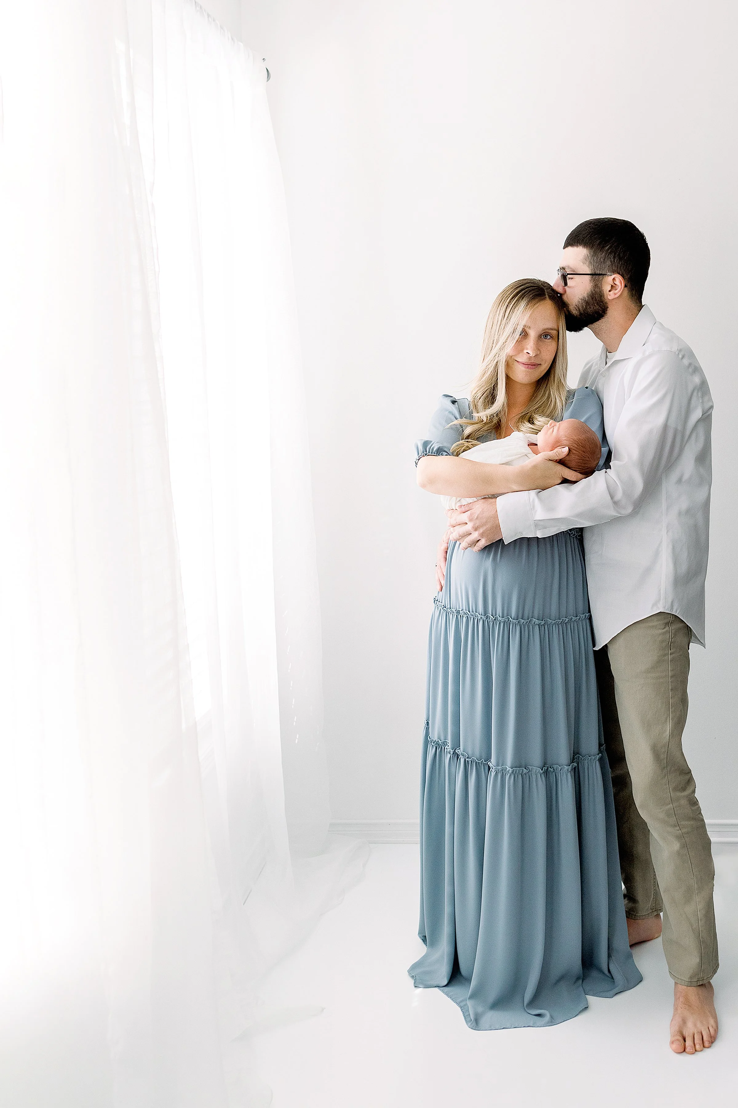 Mom in blue dress holding newborn and dad cuddling in white studio