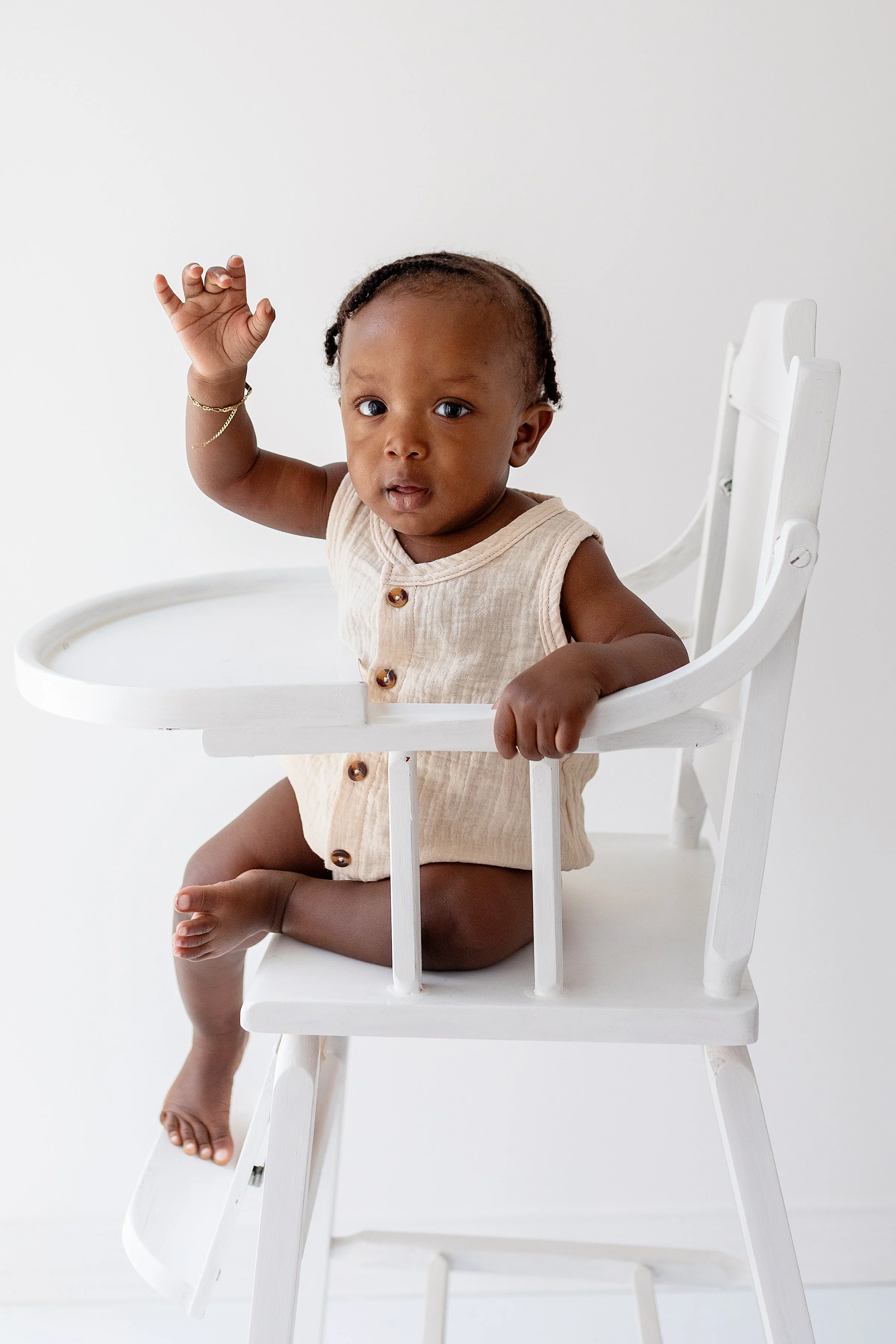 Cute one year old baby boy wearing a cream romper sitting in a white high chair waving his hand, in Oshawa baby studio