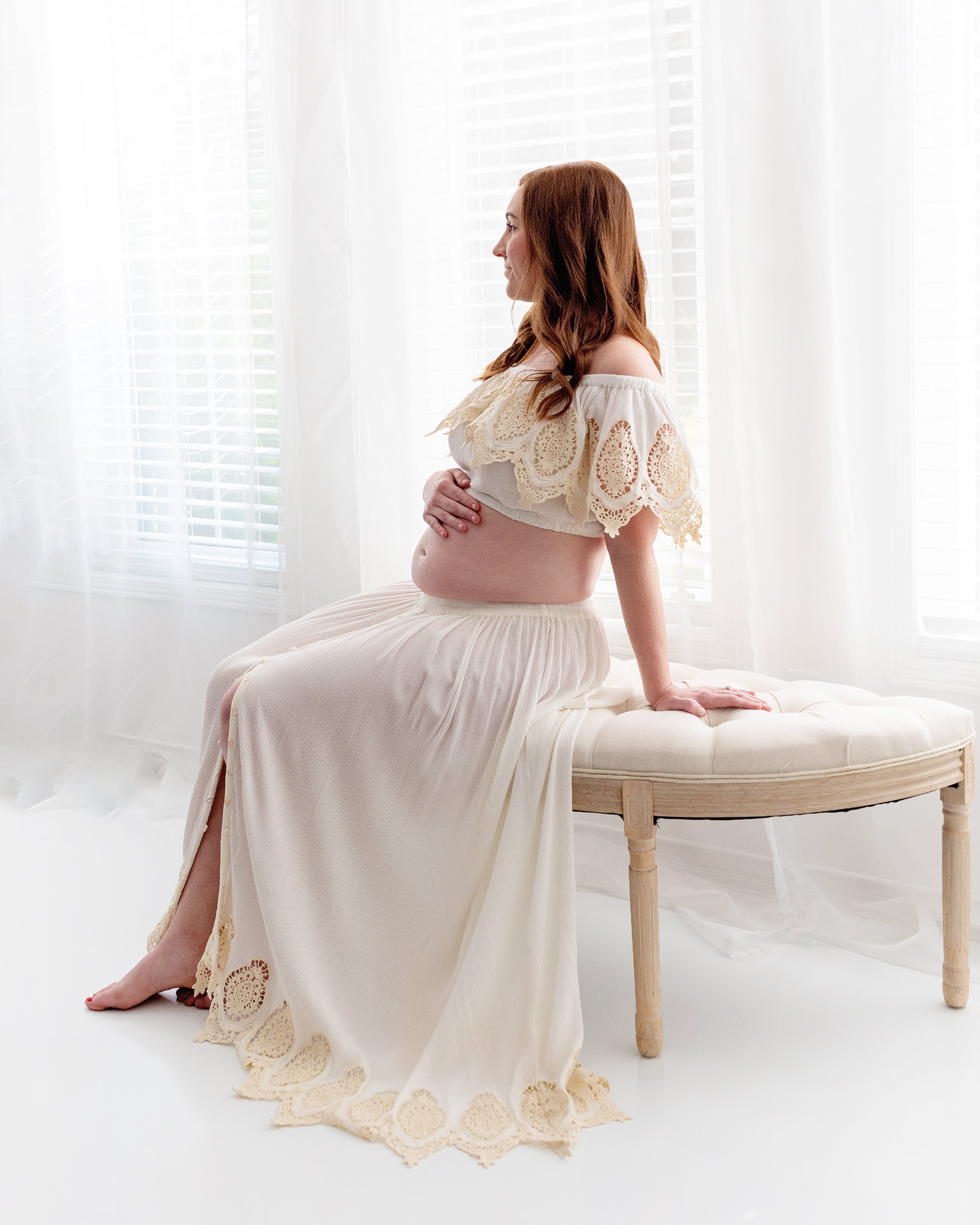 Red head mom to be in boho outfit sitting on cream bench in front of window