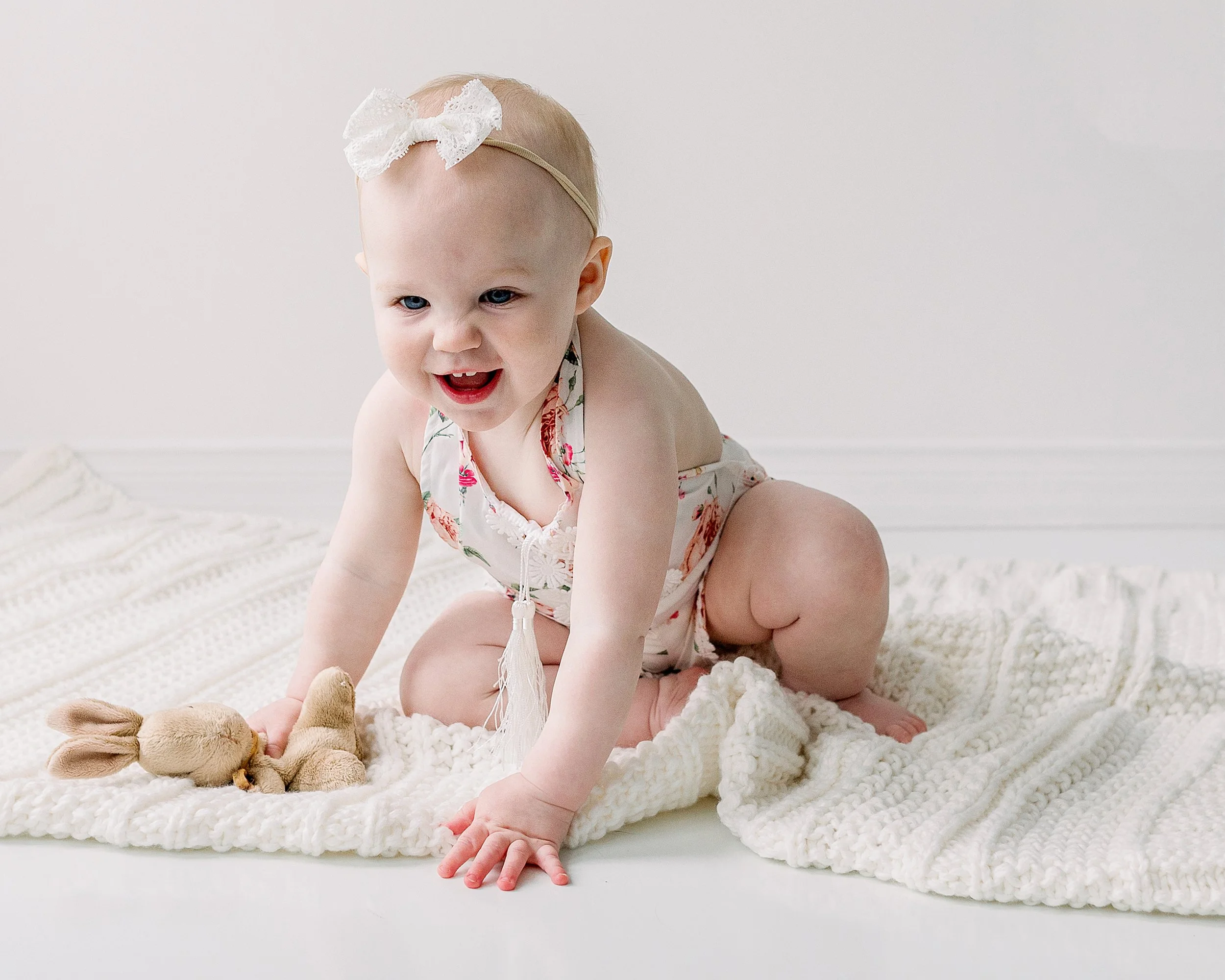 Excited one year old baby girl about to crawl off a white blanket wearing a floral romper and bow headband during baby milestone session in Oshawa