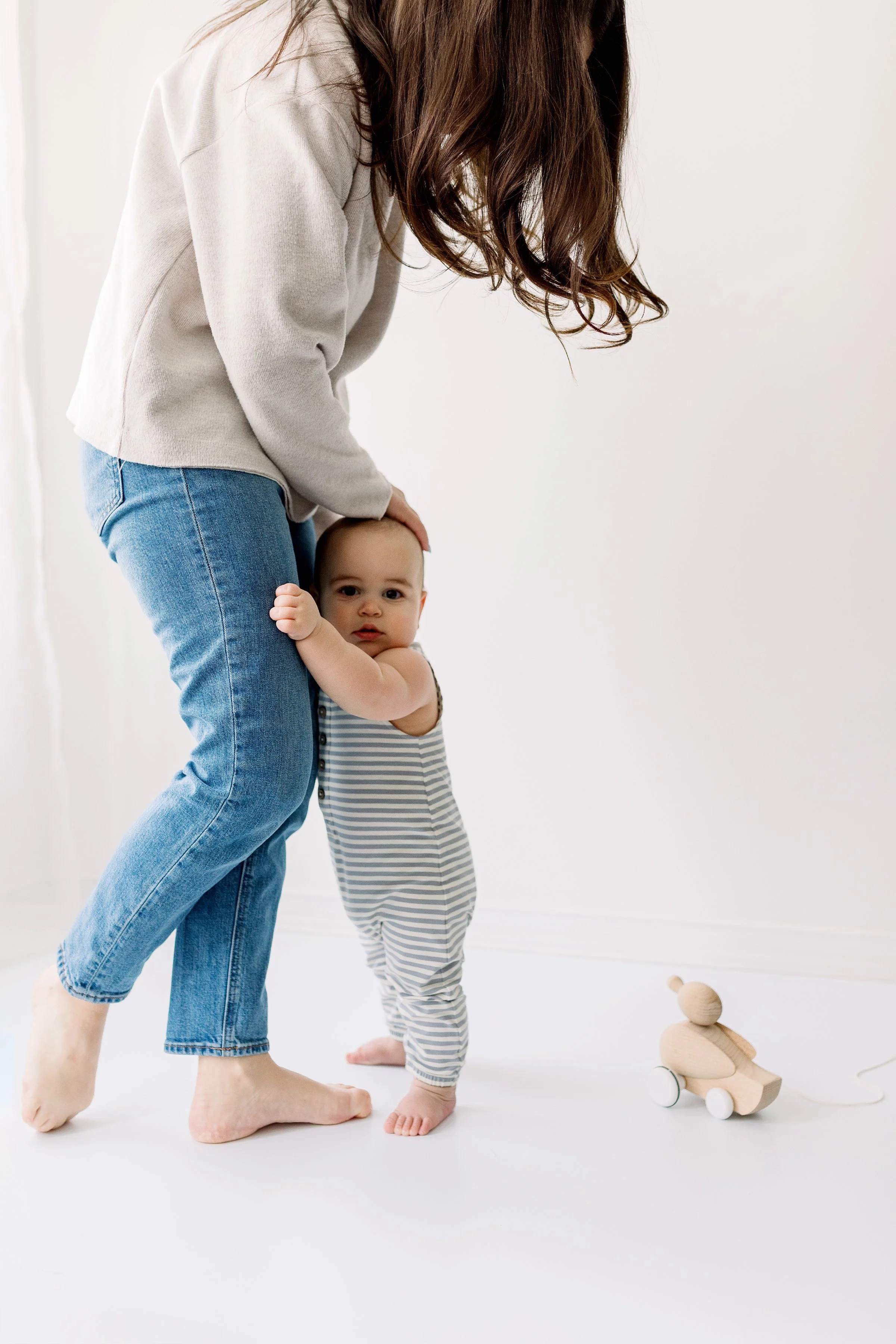 A woman holding a baby in a striped onesie standing on a white floor with a wooden toy car nearby.