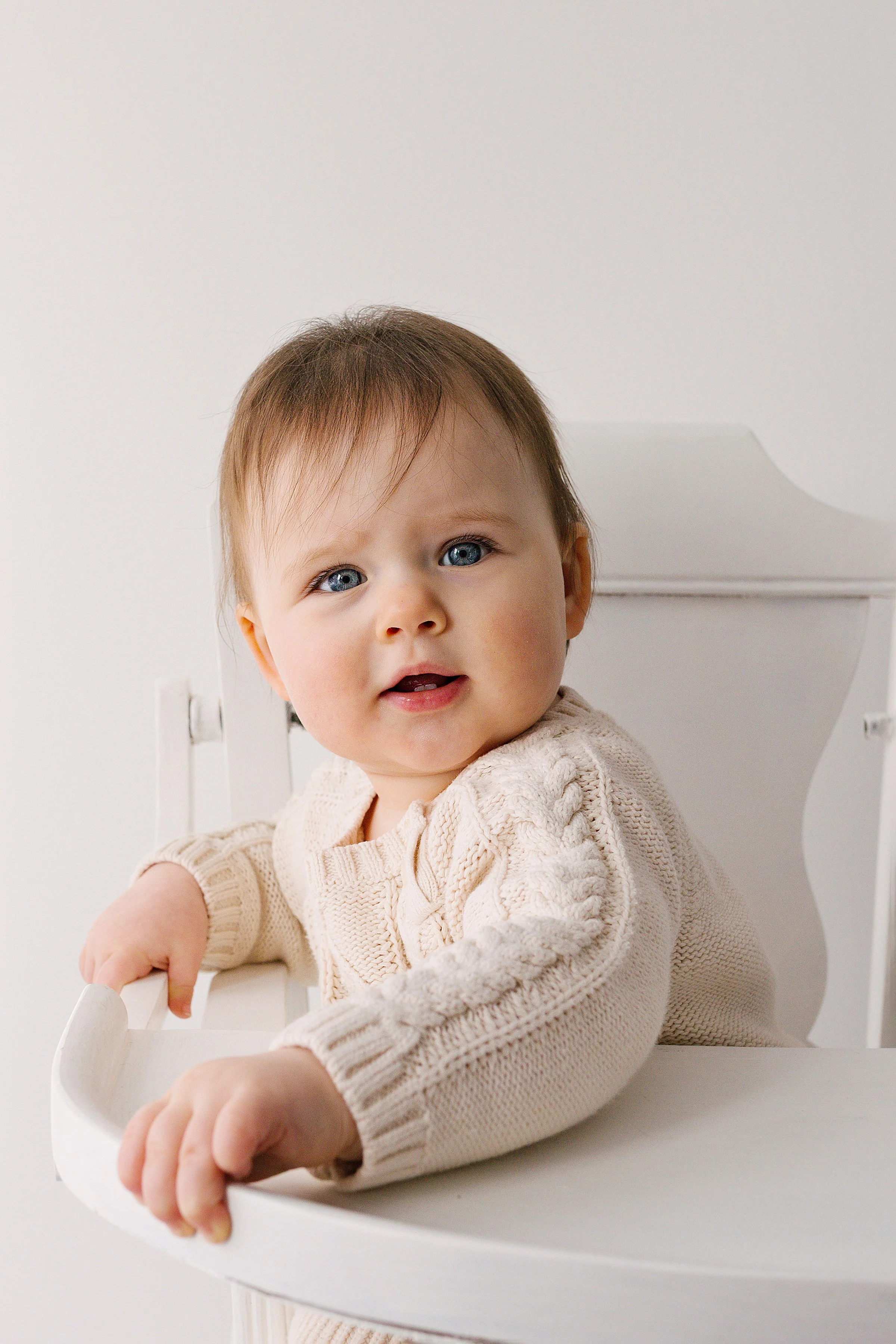 Sweet 6 month old baby girl with blue eyes and red hair sitting in a white vintage highchair wearing  knitted dress looking at the camera during baby session in Oshawa