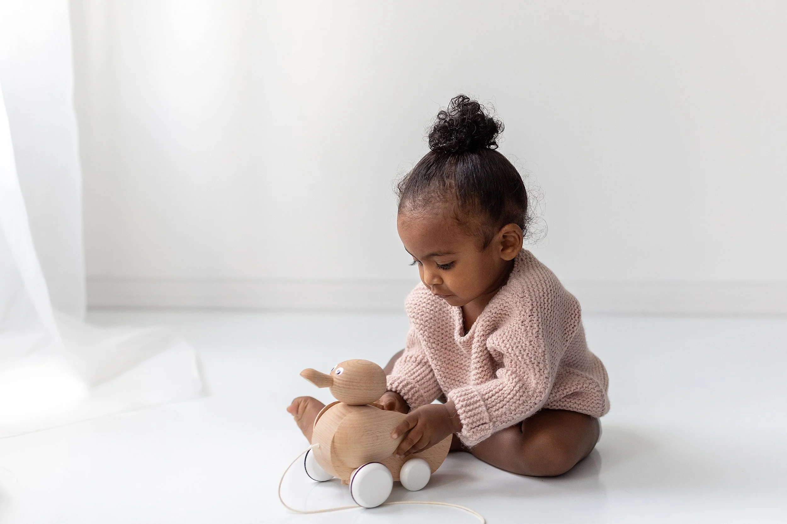 6 month old baby wearing a pink knitted sweater playing with a wooden duck on a white studio floor during baby session in Oshawa