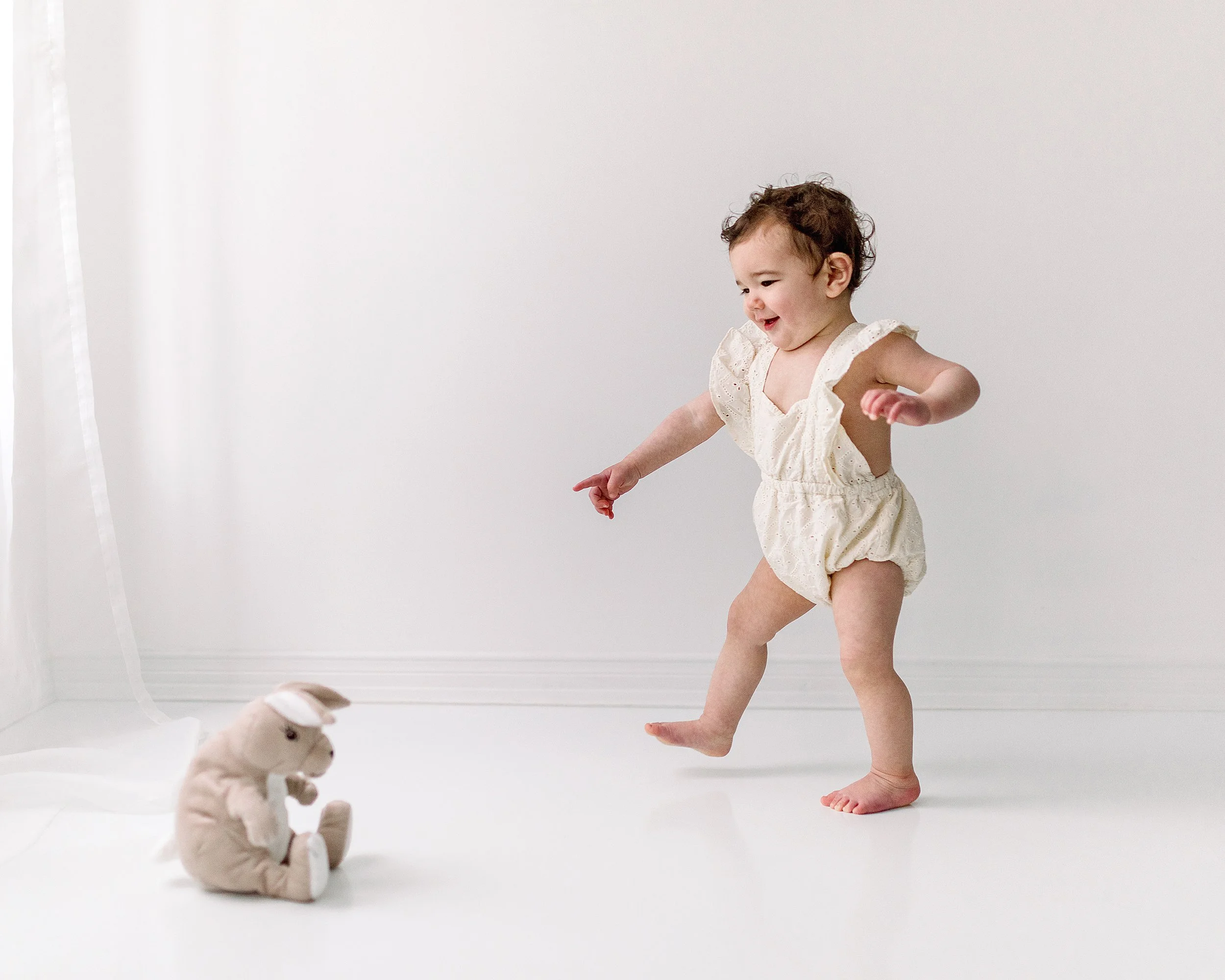 Baby girl taking steps towards a stuffed kangaroo in a white studio wearing a soft yellow baby romper smiling during baby milestone session in Durham Region