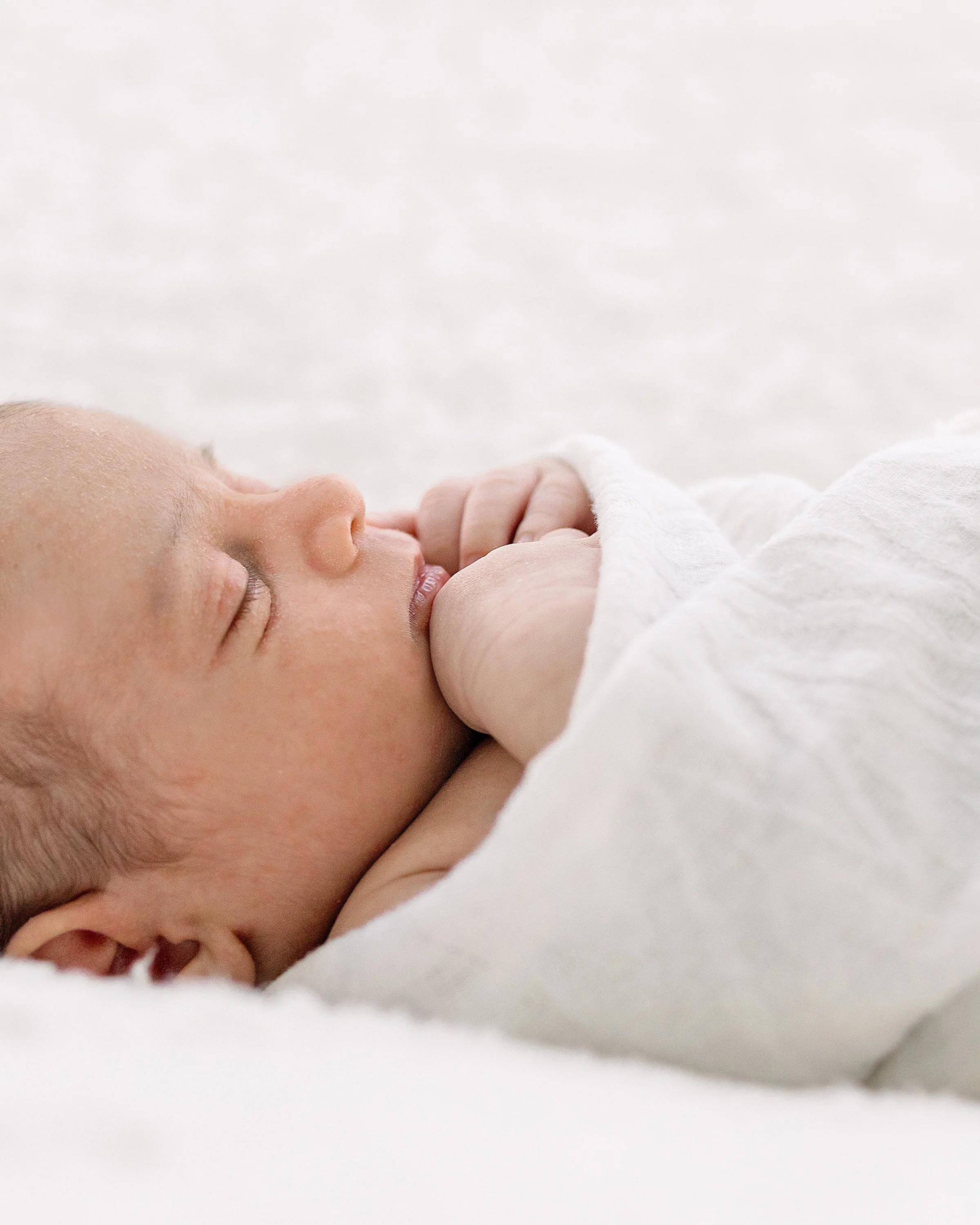Close-up of a sleeping baby with hands near face, resting on a soft white surface.