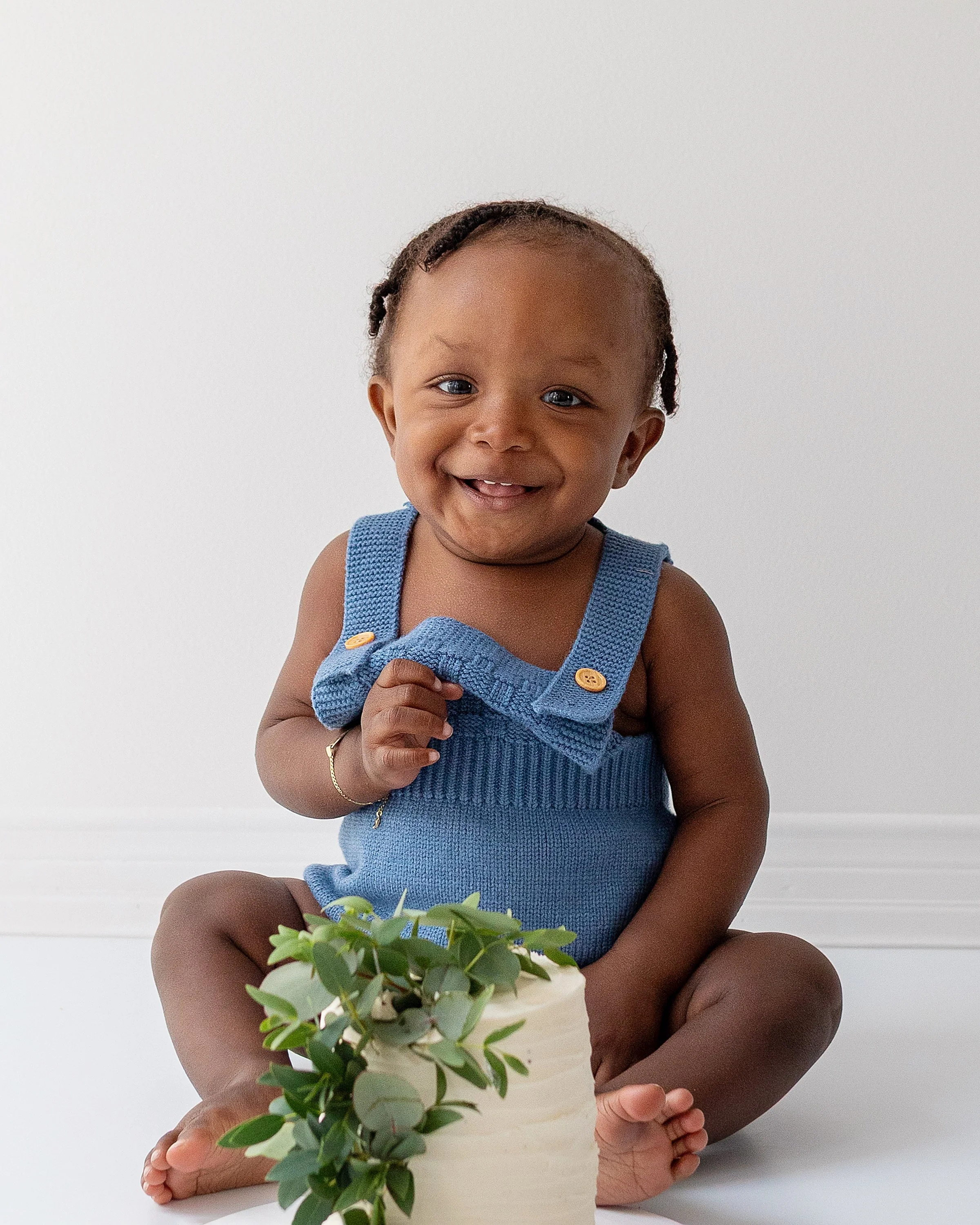 Smiling baby boy sitting on the floor with his white cake and greenery wearing a blue romper, in Oshawa studio