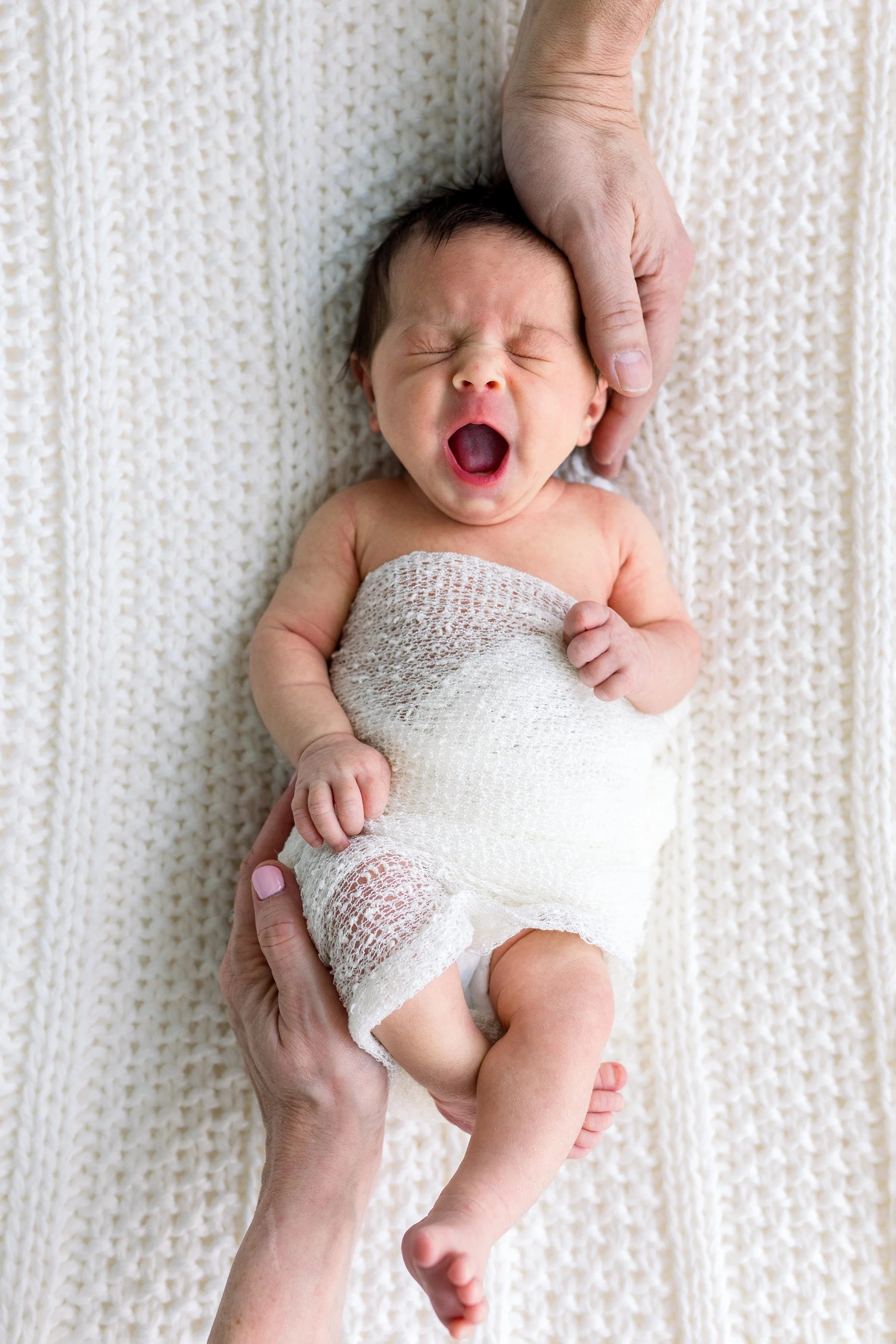 Yawning Newborn with mom and dad holding head and feet