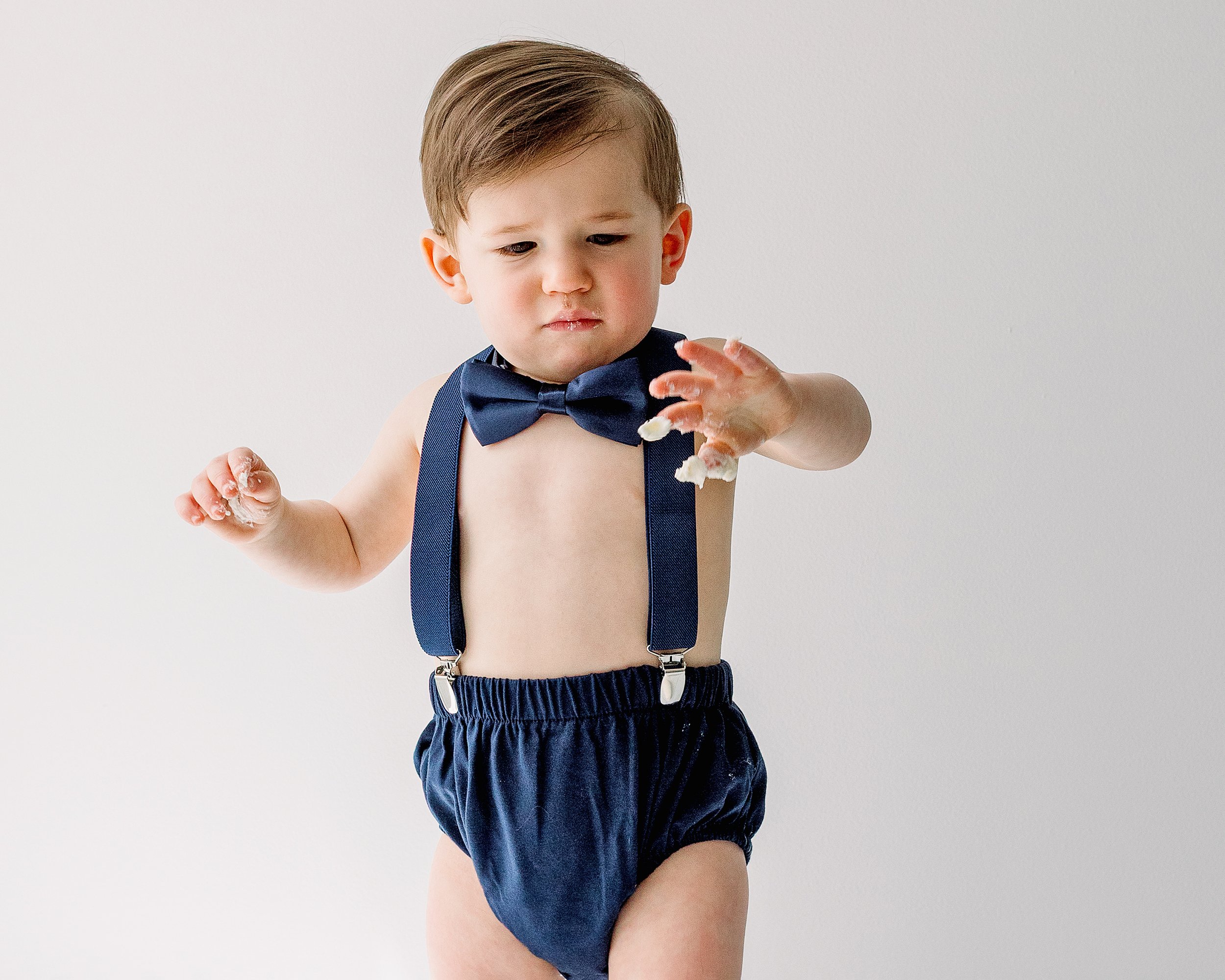 Baby in navy blue romper and bowtie shaking icing of his fingers in a white studio, in Oshawa