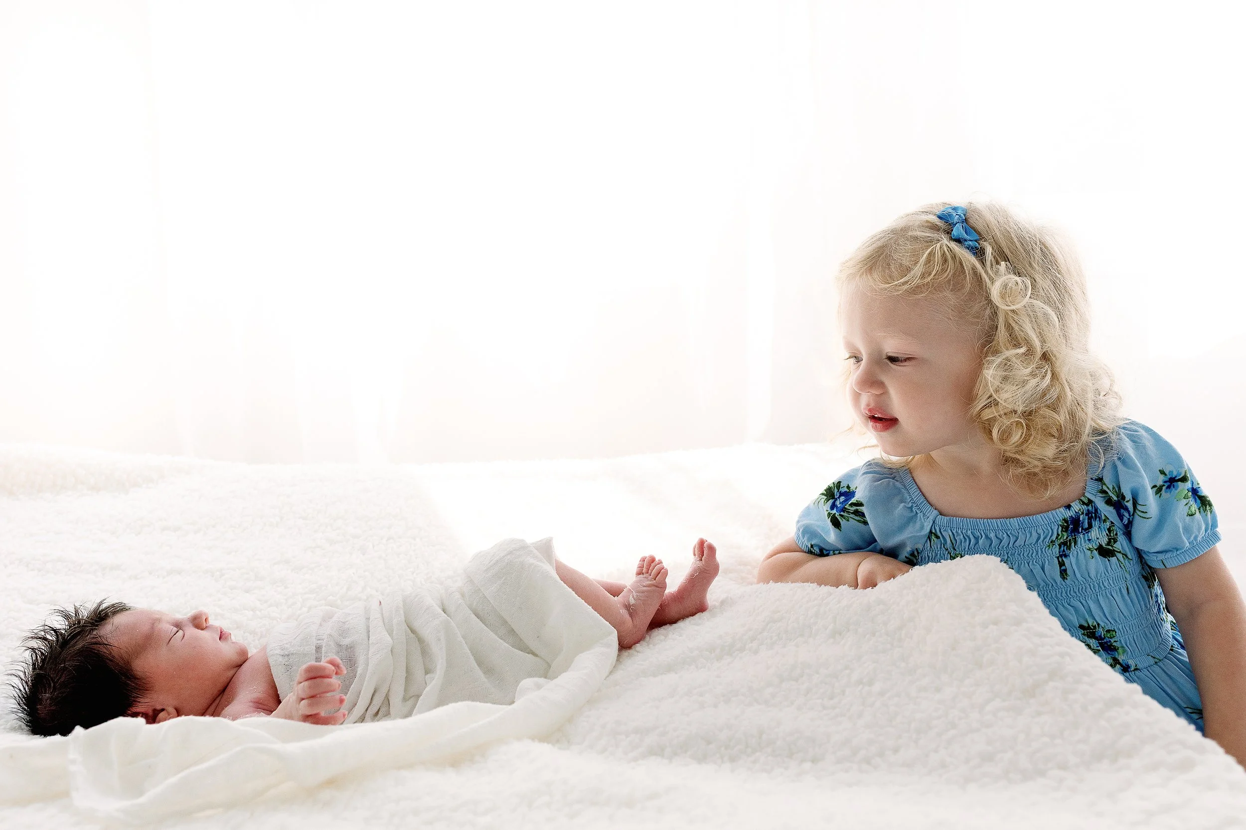 A young girl with curly blonde hair and wearing a blue dress with floral embroidery is lying on her stomach on a white bed, looking at a tiny newborn baby wrapped in a white blanket.
