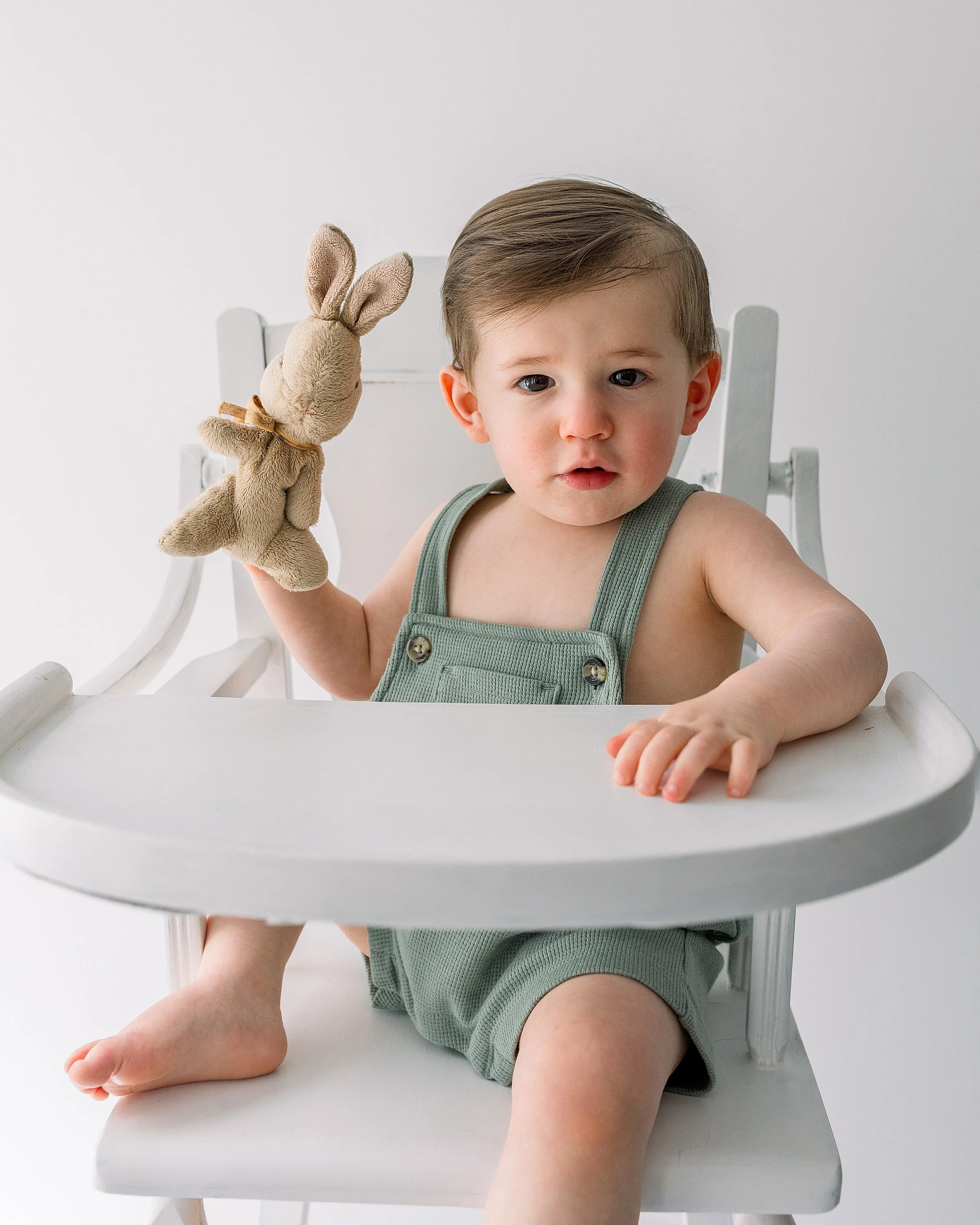 Handsome one year old  boy in olive green romper sitting in white vintage high chair looking at camera holding a stuffed bunny, in Oshawa baby studio