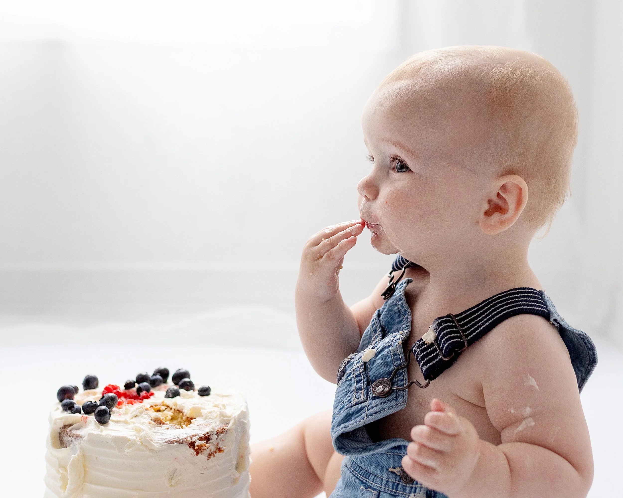 Blonde baby boy in blue overalls sitting on the floor eating cake looking straight ahead in front of white window, in Oshawa