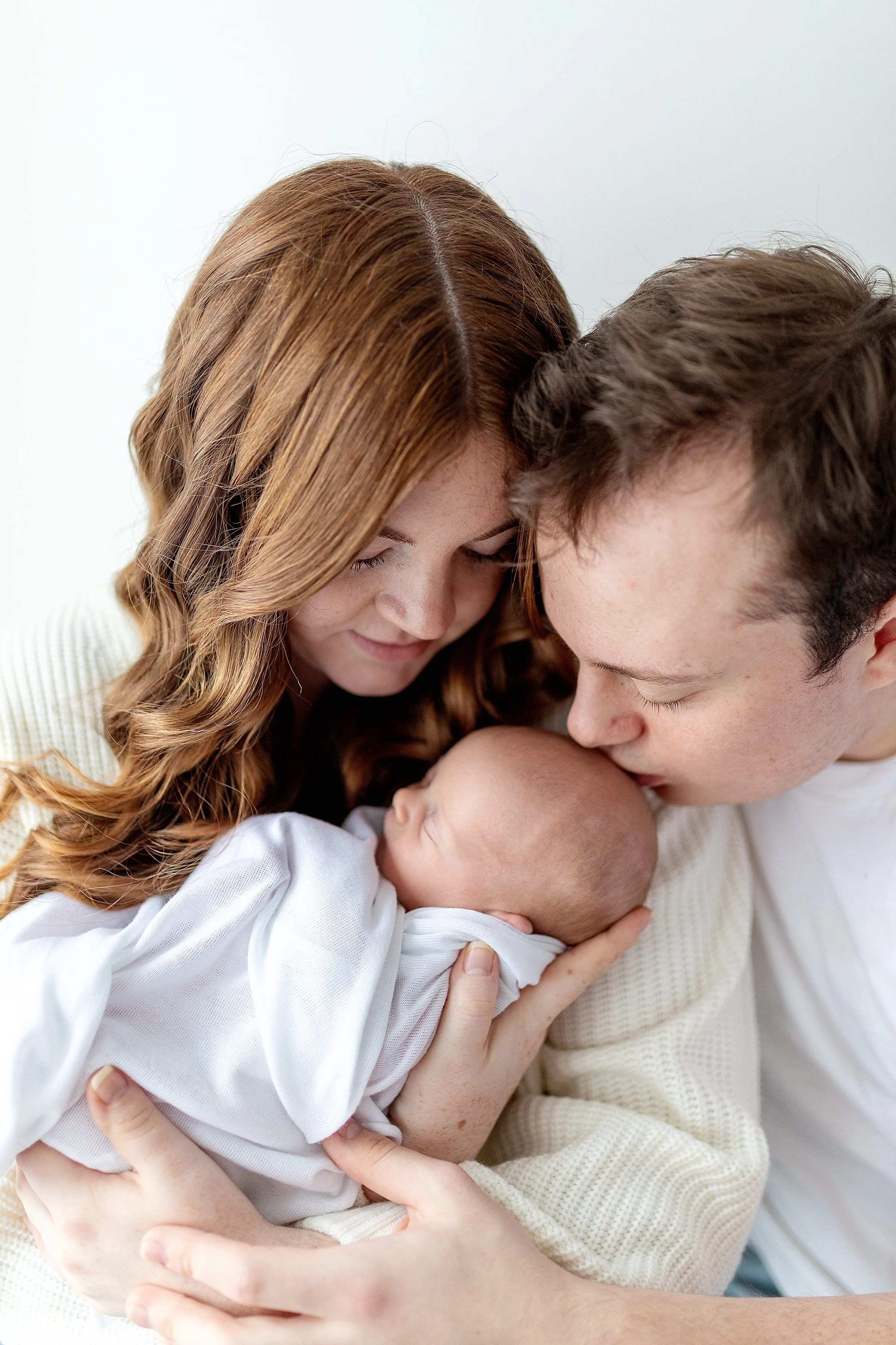 Young new parents looking down at their newborn baby girl in white wrap smiling and kissing her head, in Durham Region