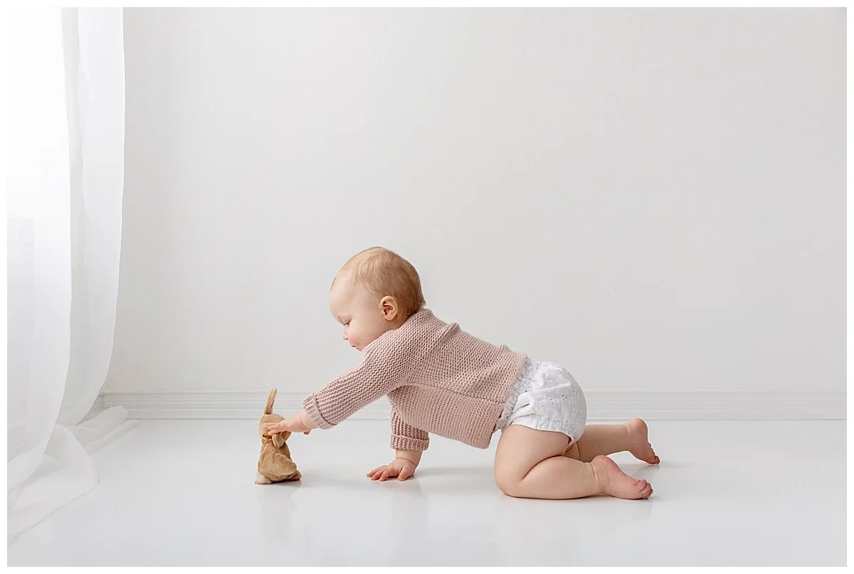 Cute baby girl crawling to a baby bunny wearing a pink knit sweater and white diaper cover in a white studio during a baby session in Oshawa