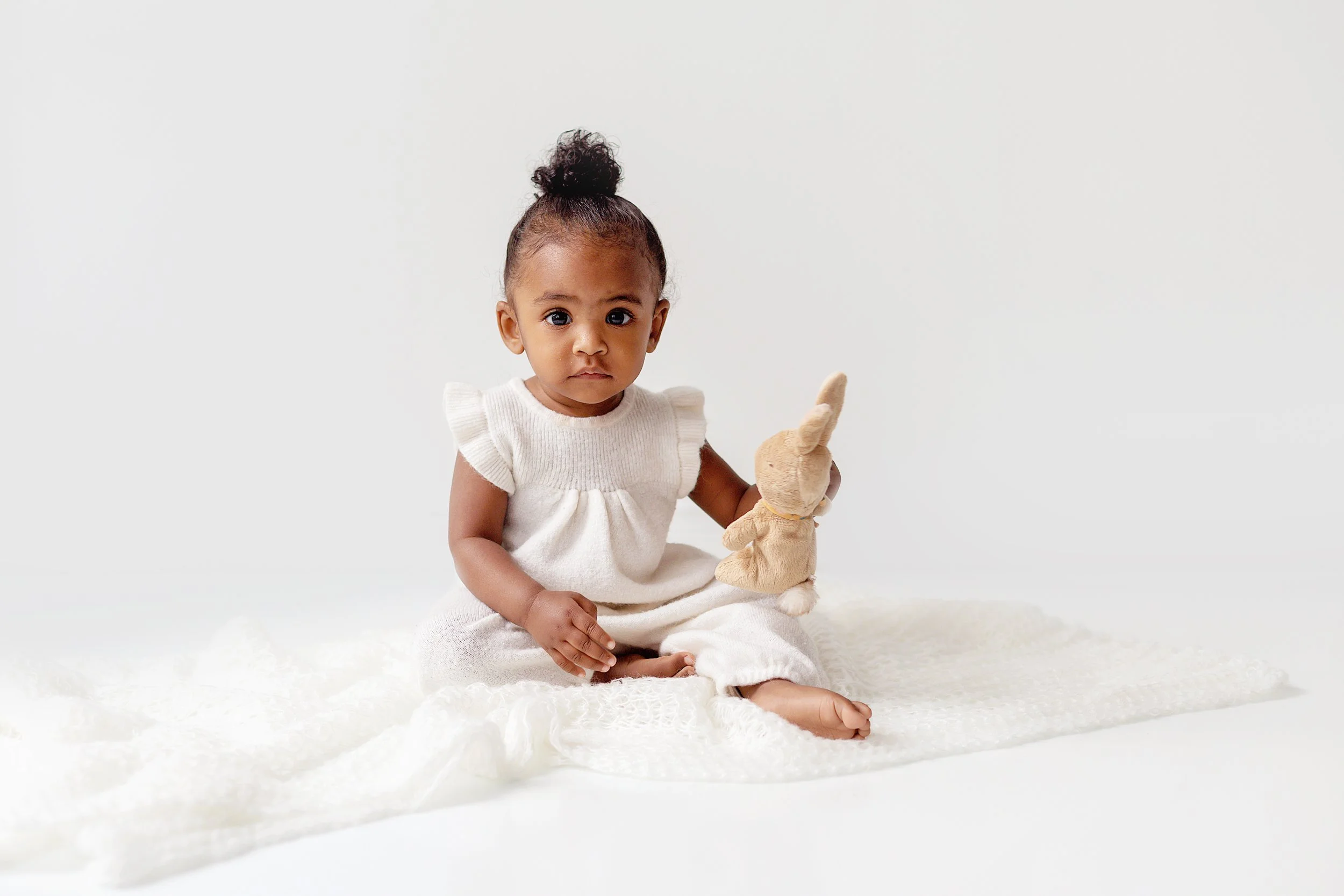 Dark skinned baby girl sitting on a white blanket wearing a white romper holding a stuffed bunny looking at the camera during baby session in Oshawa