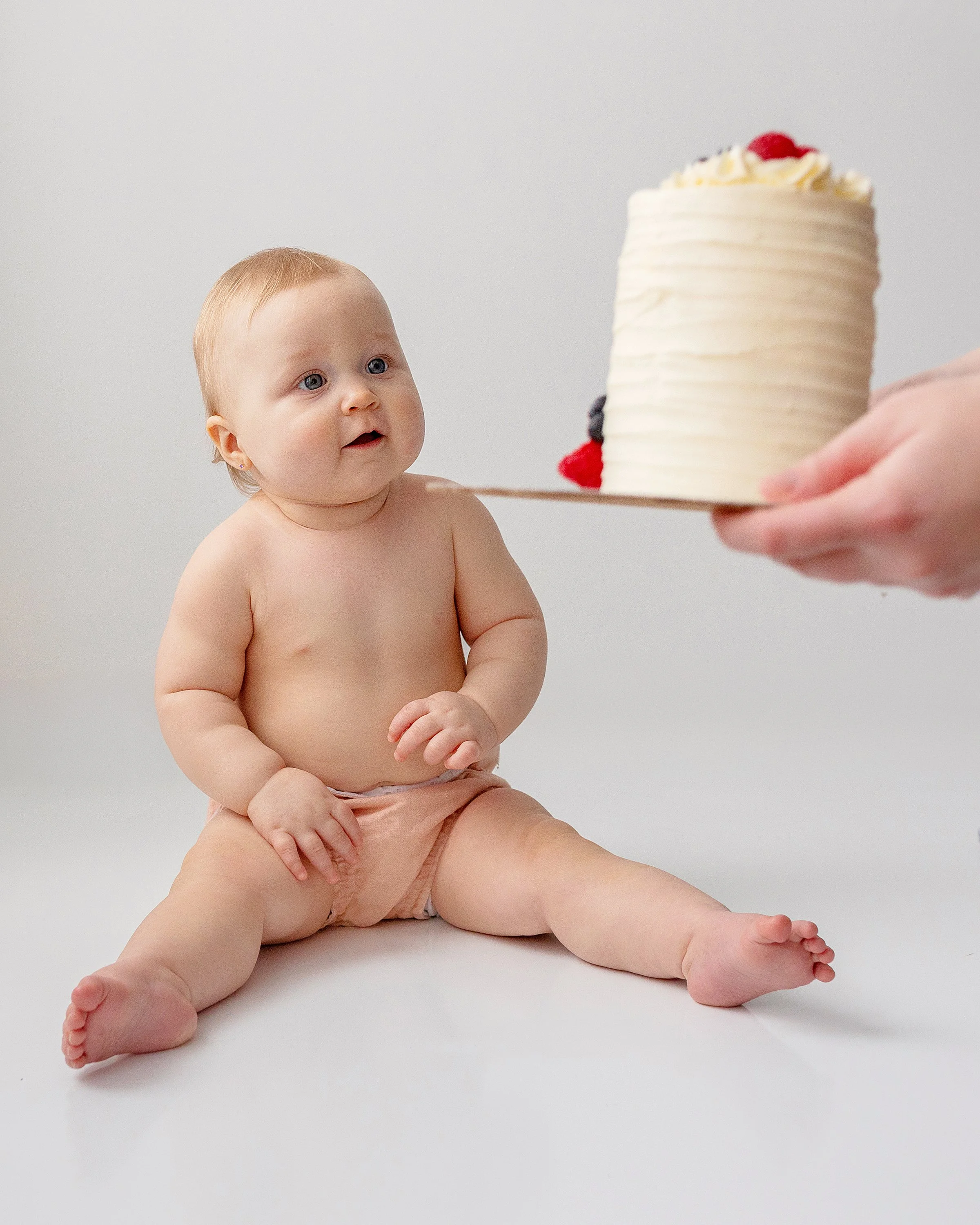 Chubby baby girl excited for her white cake and fresh berries that mom is holding in front of her, in Oshawa studio