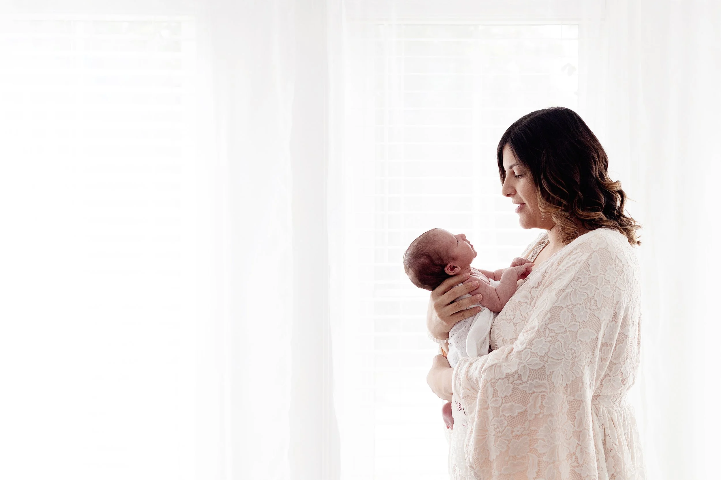 New mom holding newborn looking at each other in front of white window