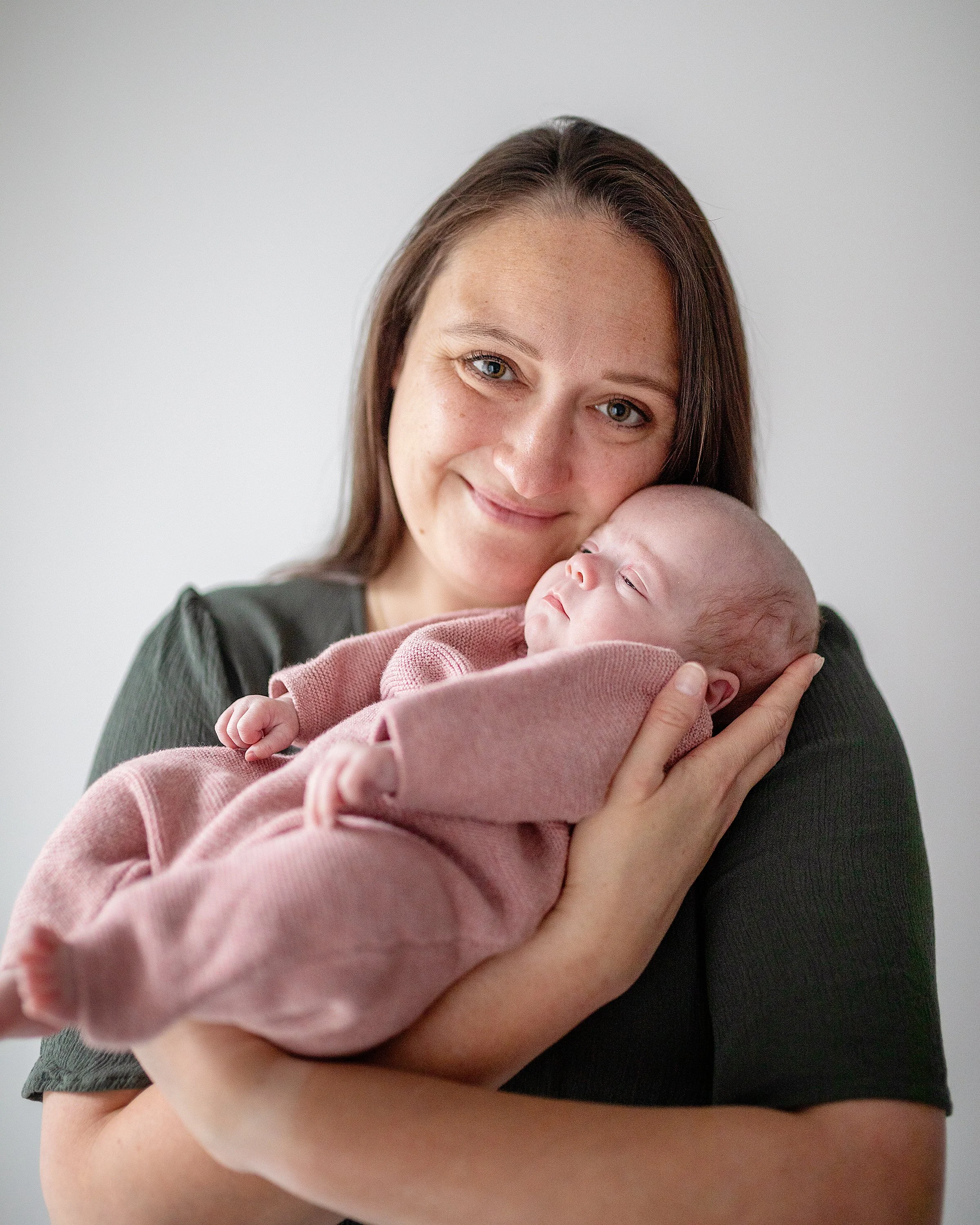 Smiling new mom holding newborn daughter in pink knitted outfit while baby sleeps in Oshawa studio