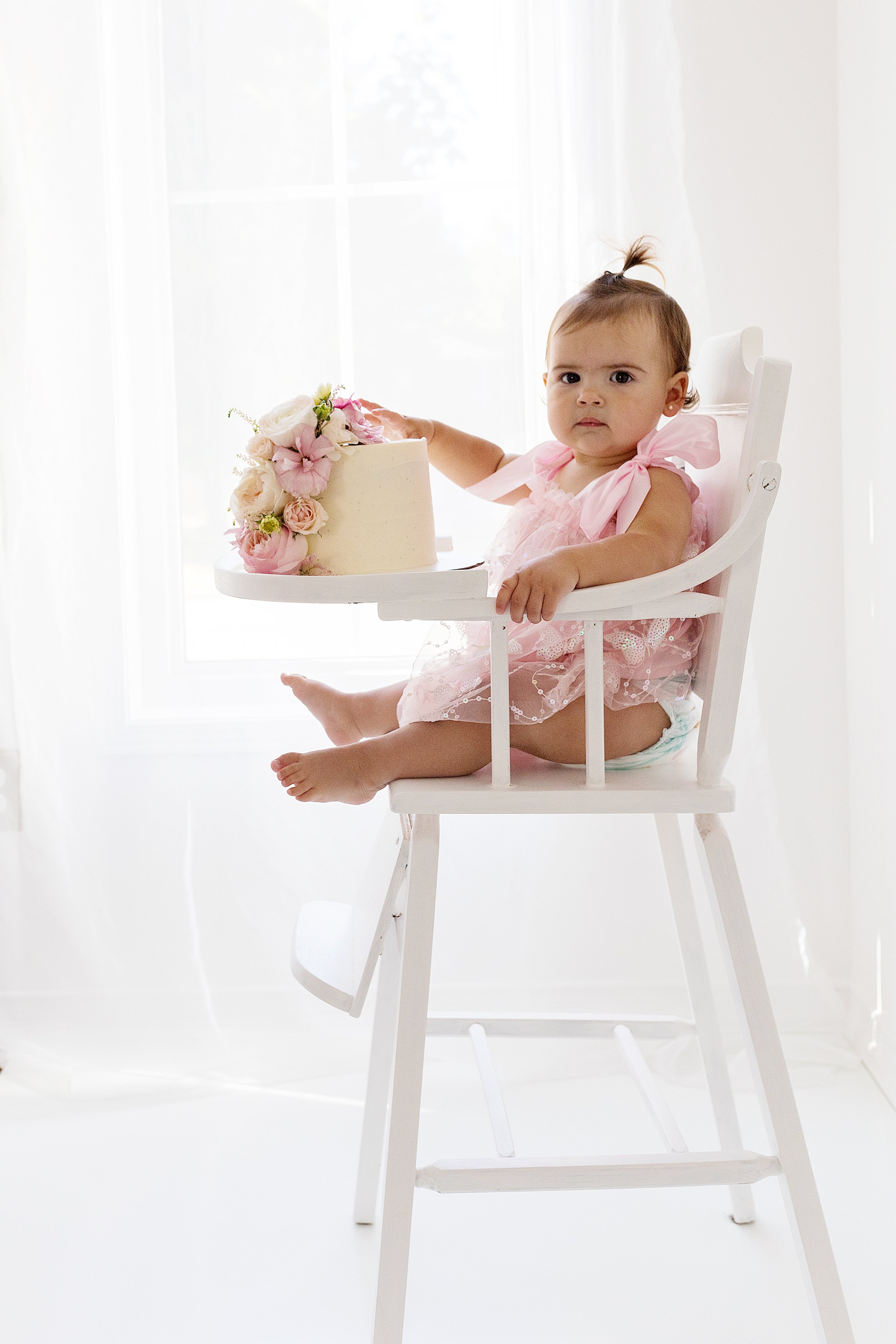 One year old baby girl looking at camera with one hand on her floral cake sitting in a vintage white highchair, in front of  a window, in Durham Region