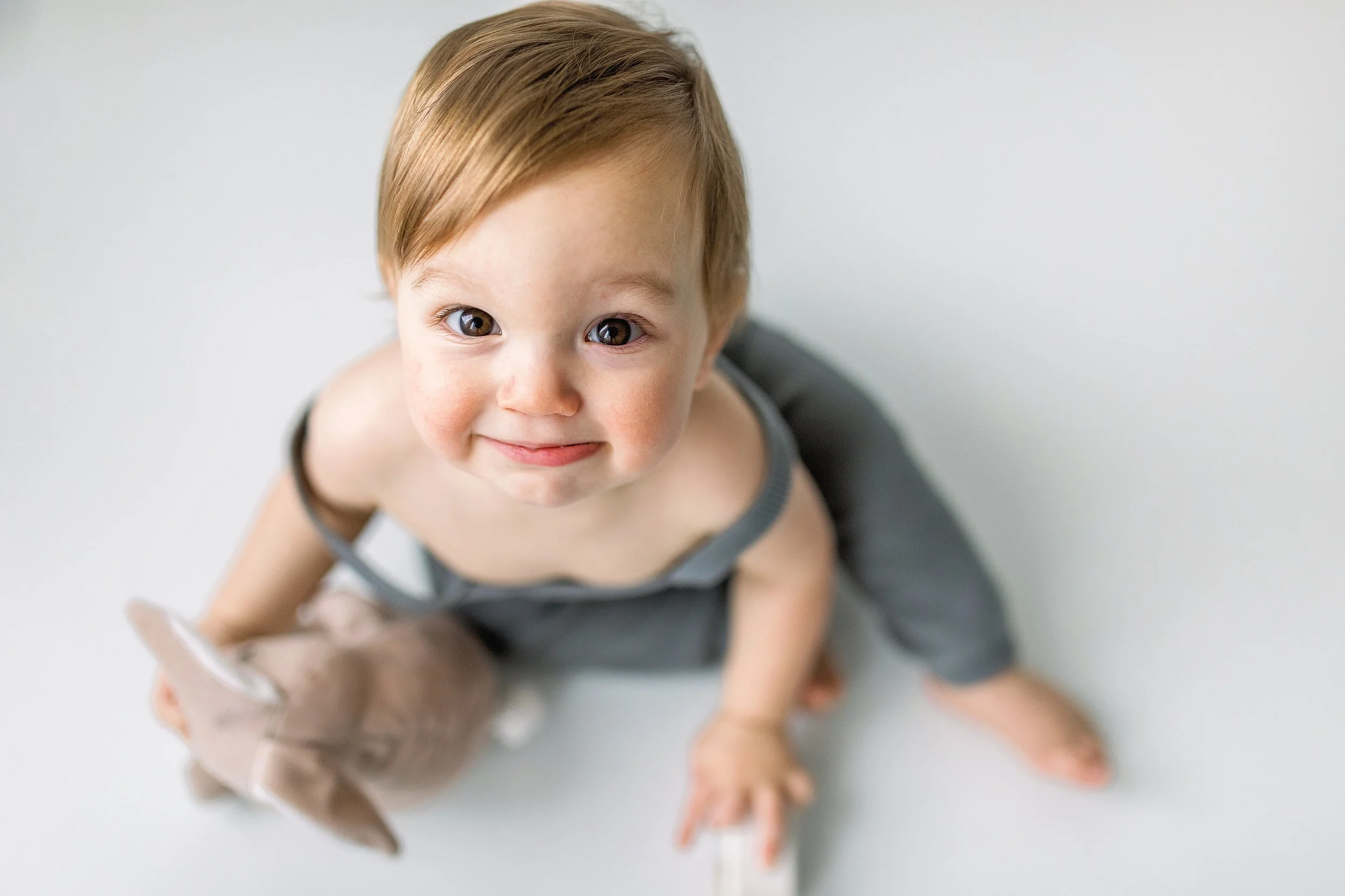 Cute baby boy with strawberry blonde hair smiling at the camera wearing a teal knit romper and holding a stuffed kangaroo during a baby session in Oshawa