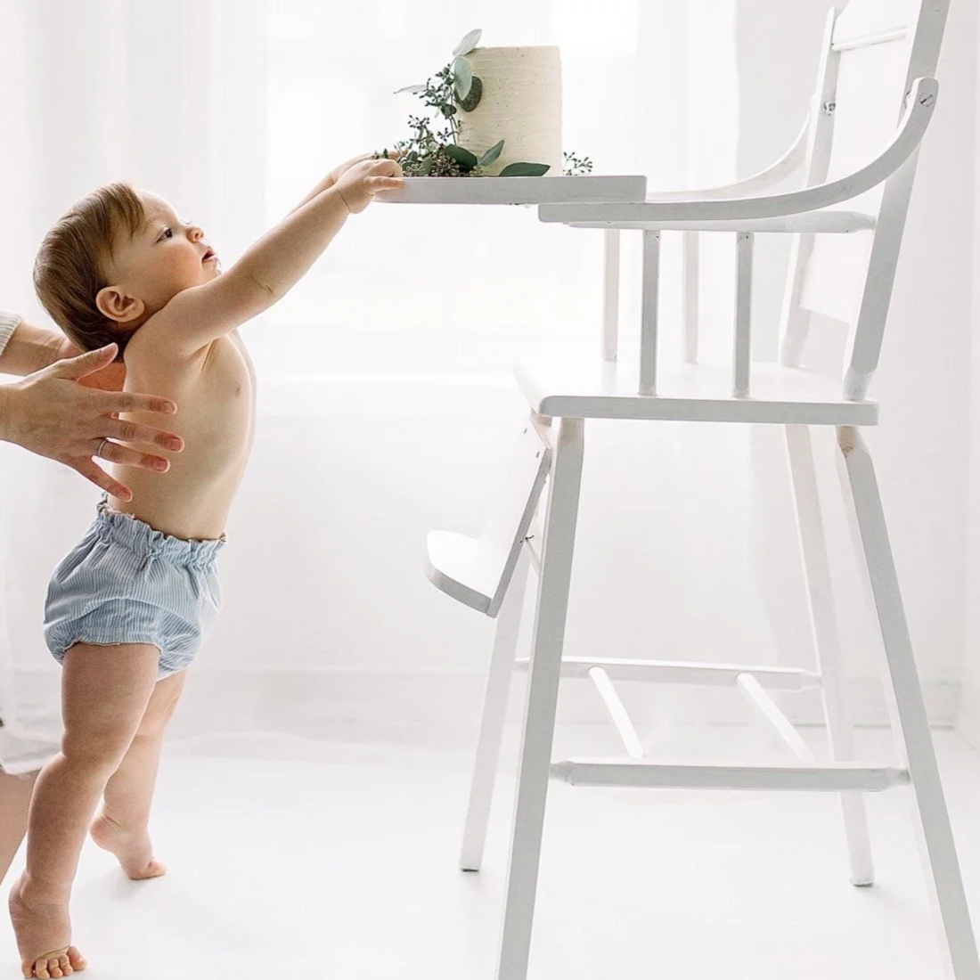A young boy in only a diaper reaching for a small white table with a plant on it, with assistance from an adult hand.