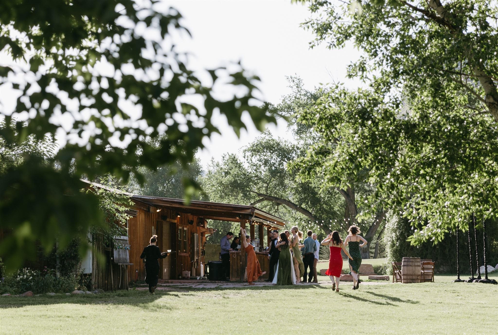 People gathered outdoors at a wedding at Planet Bluegrass under trees next to a wooden building, with some dancing and socializing.
